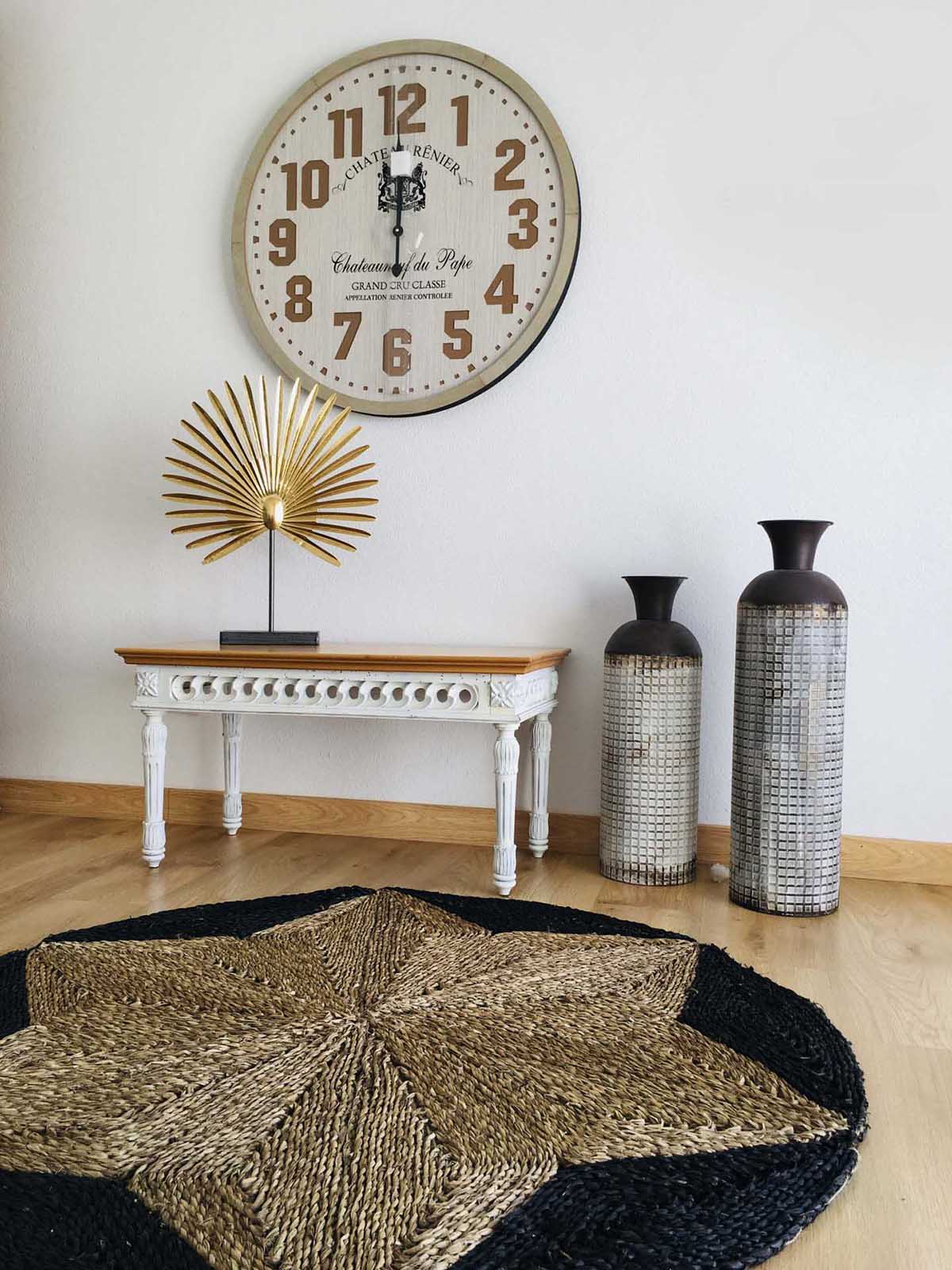 Relógio de parede redondo com números castanhos, mesa branca pequena, decoração dourada, dois vasos prateados e tapete redondo em padrão de estrela