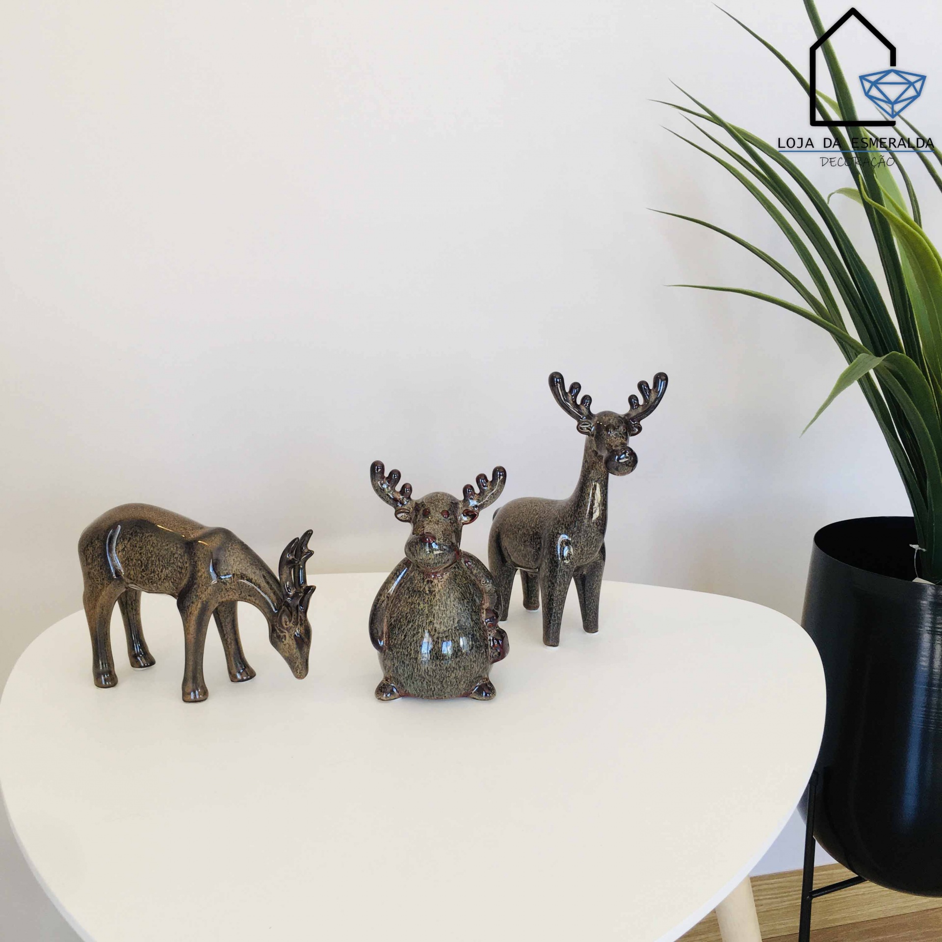 Três esculturas decorativas de veados em metal castanho numa mesa branca ao lado de uma planta