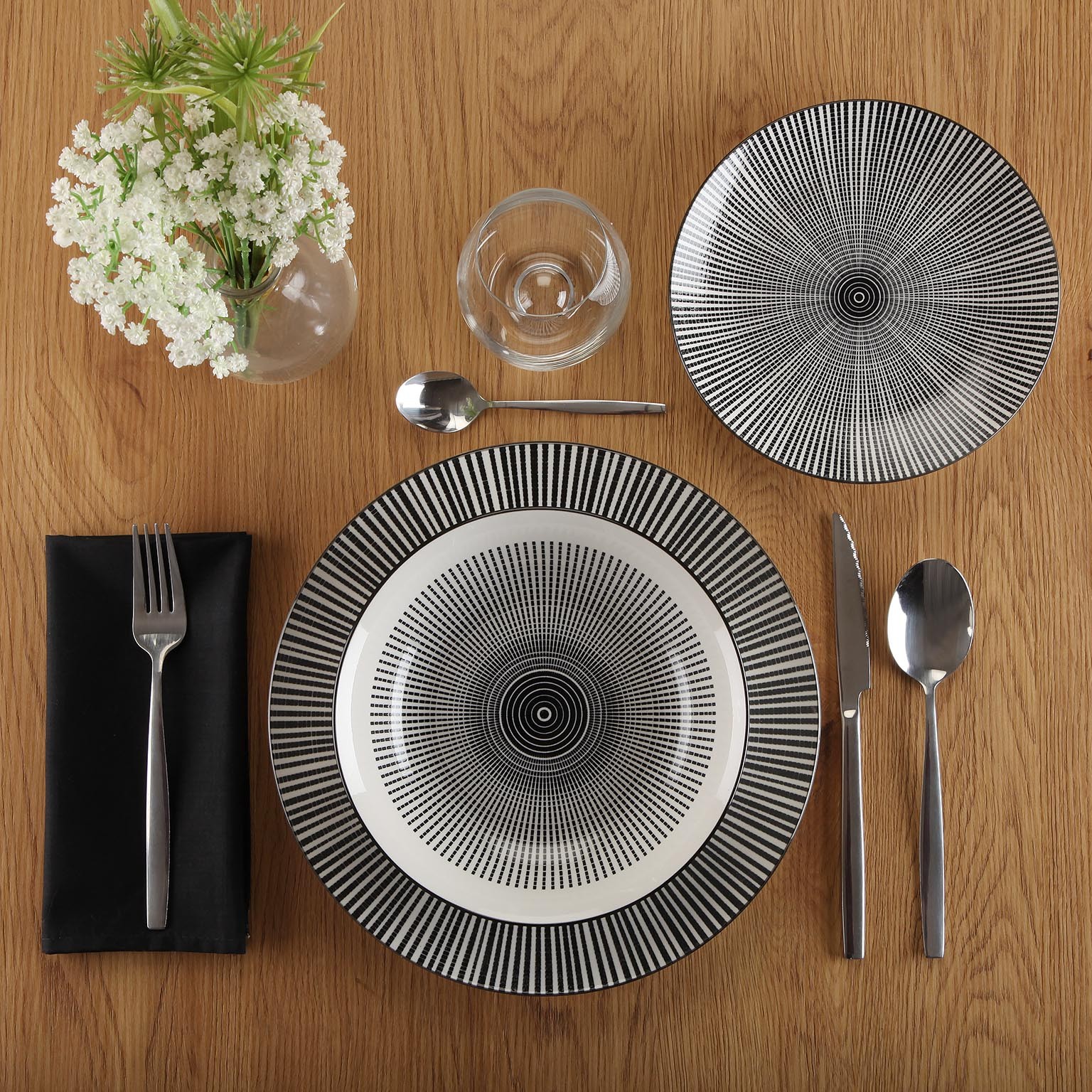 Conjunto de loiça preto e branco com talheres e copo numa mesa de madeira clara