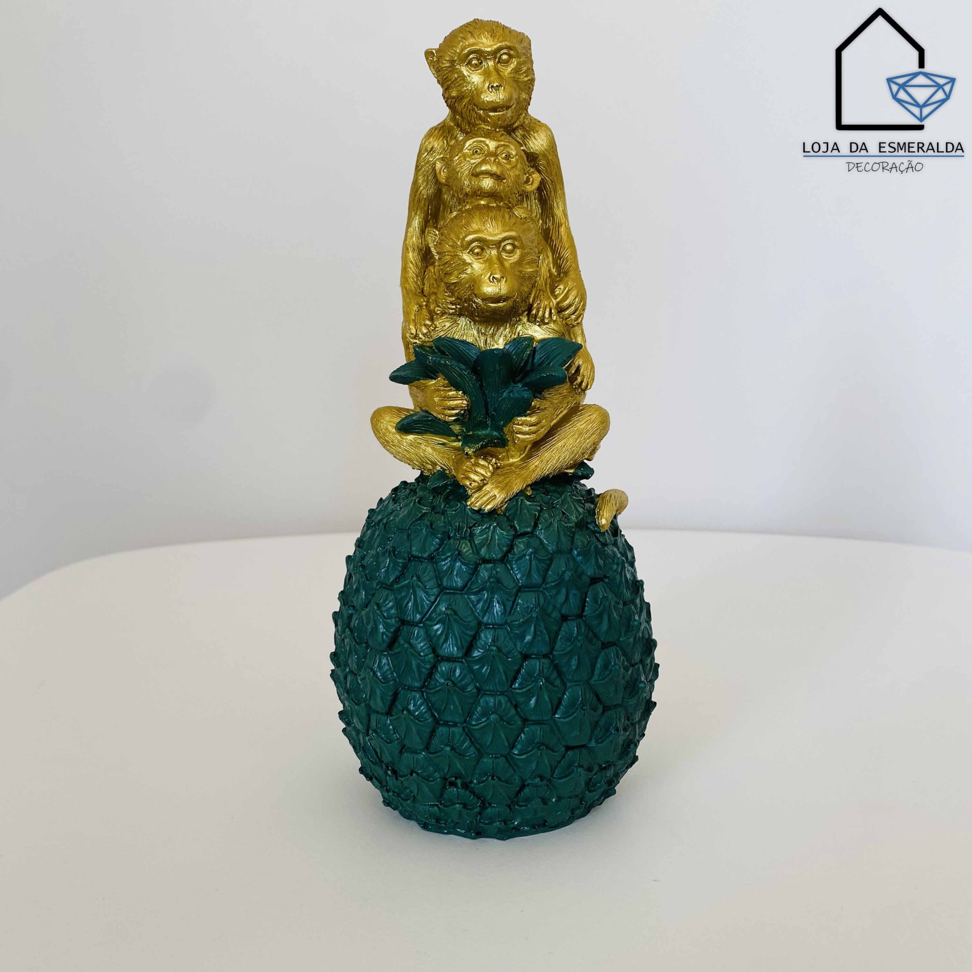 Estatueta de três macacos dourados sobre pinha azul-esverdeada em fundo branco