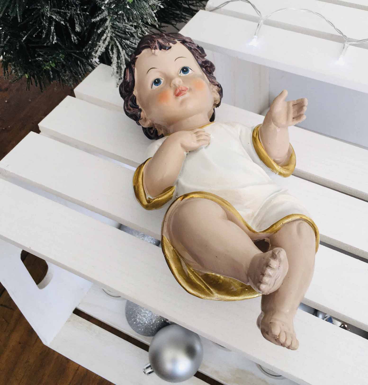 Estátua do bebé Jesus em cerâmica com túnica branca e detalhes dourados, sobre suporte branco com bolas prateadas.