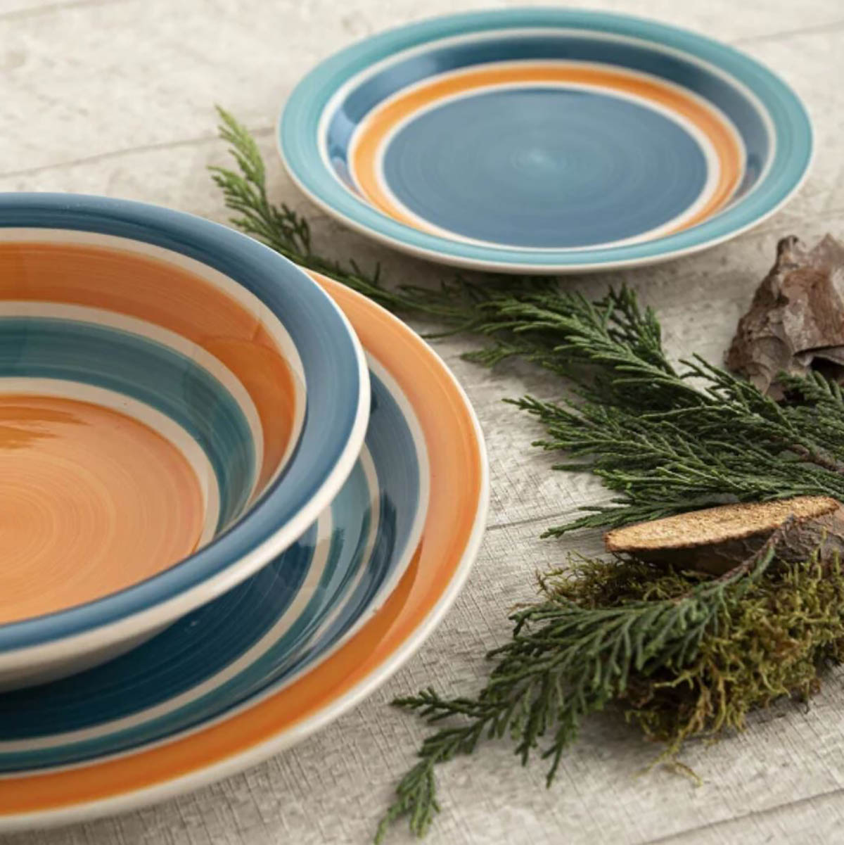 Conjunto de pratos e tigelas em cerâmica com listras azul, laranja e bege sobre superfície branca com ramos verdes e cascas.