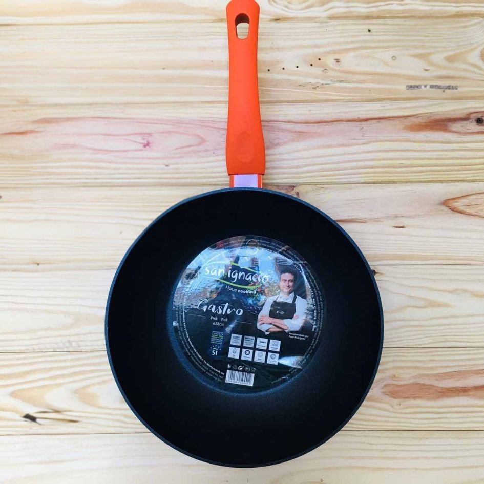 Wok preto com cabo laranja e etiqueta redonda em superfície de madeira.