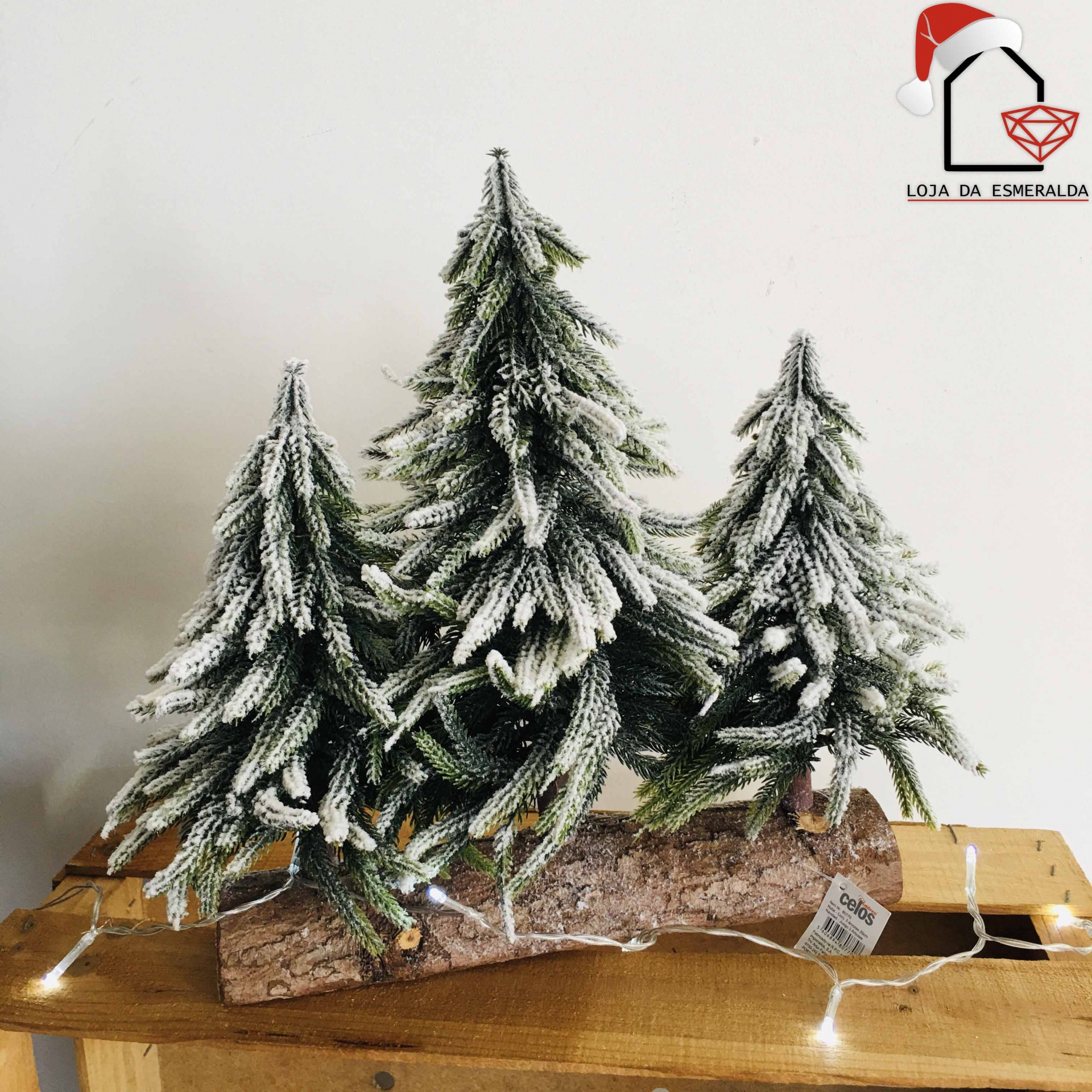 Três árvores de Natal artificiais com neve sobre tronco de madeira numa mesa de madeira clara