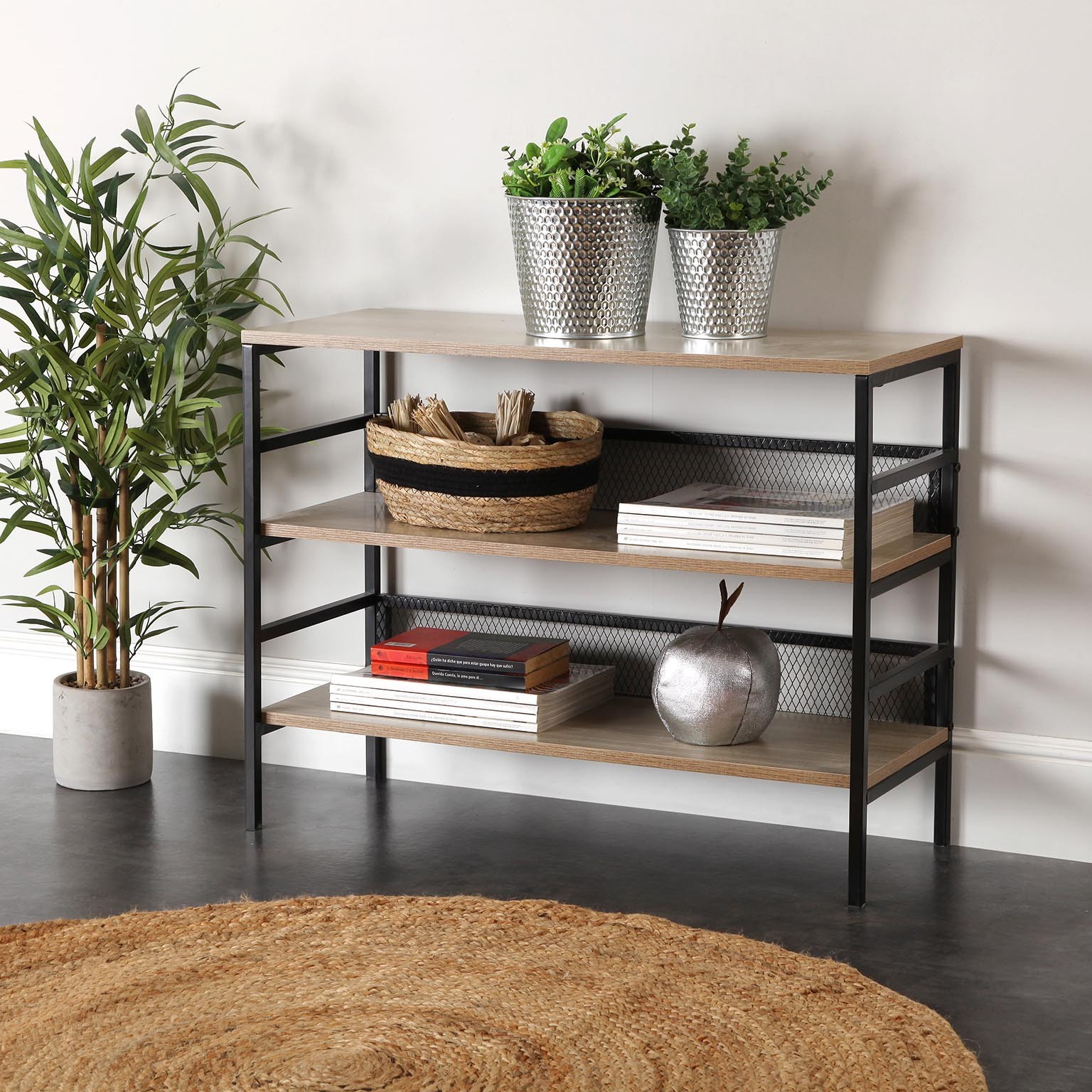 Estante de madeira clara com estrutura metálica preta, vasos prateados com plantas, cesta de palha e livros, em ambiente interior com tapete redondo de fibra natural.