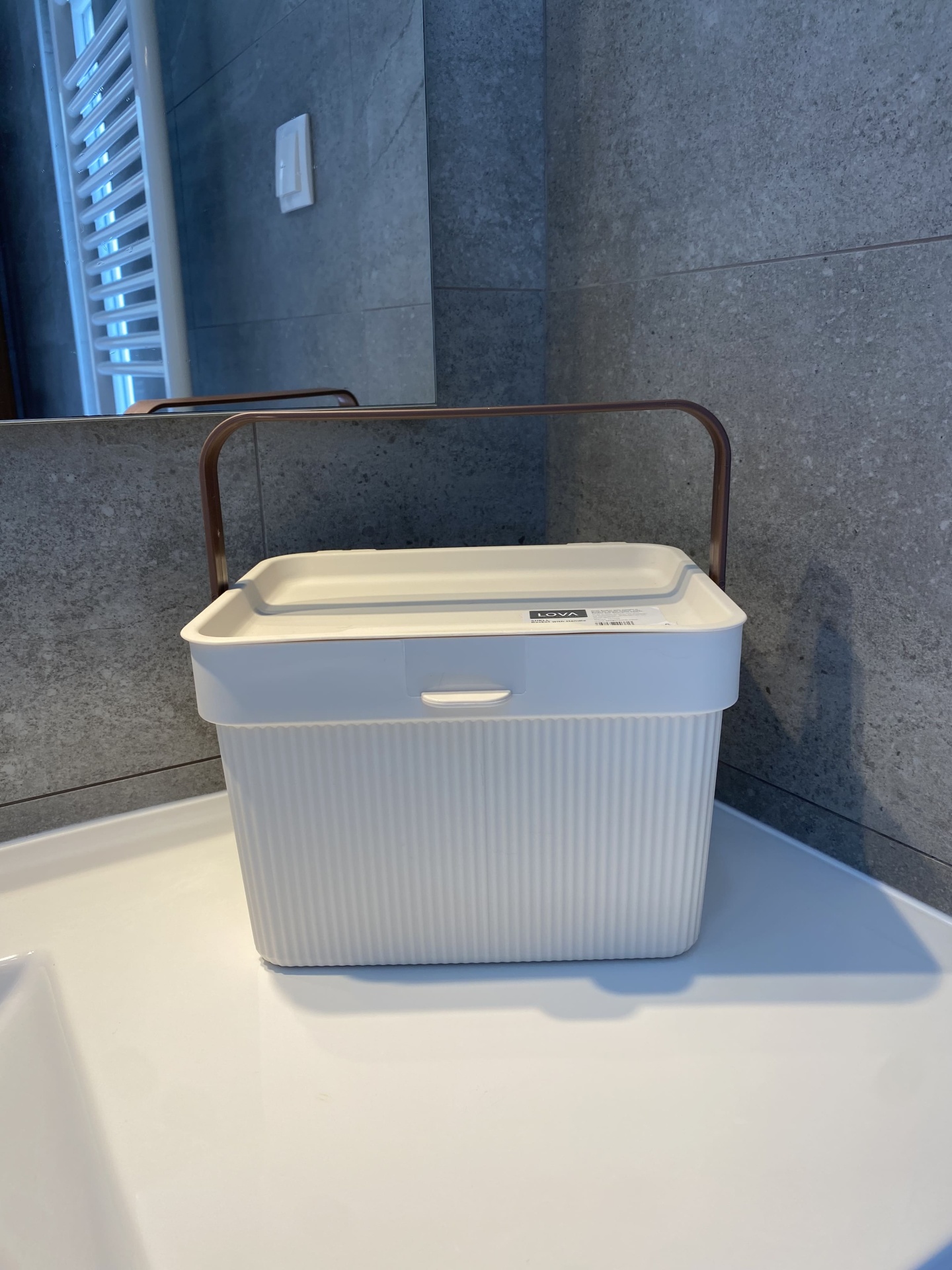 Caixa de armazenamento branca em plástico com tampa e pega castanha numa casa de banho