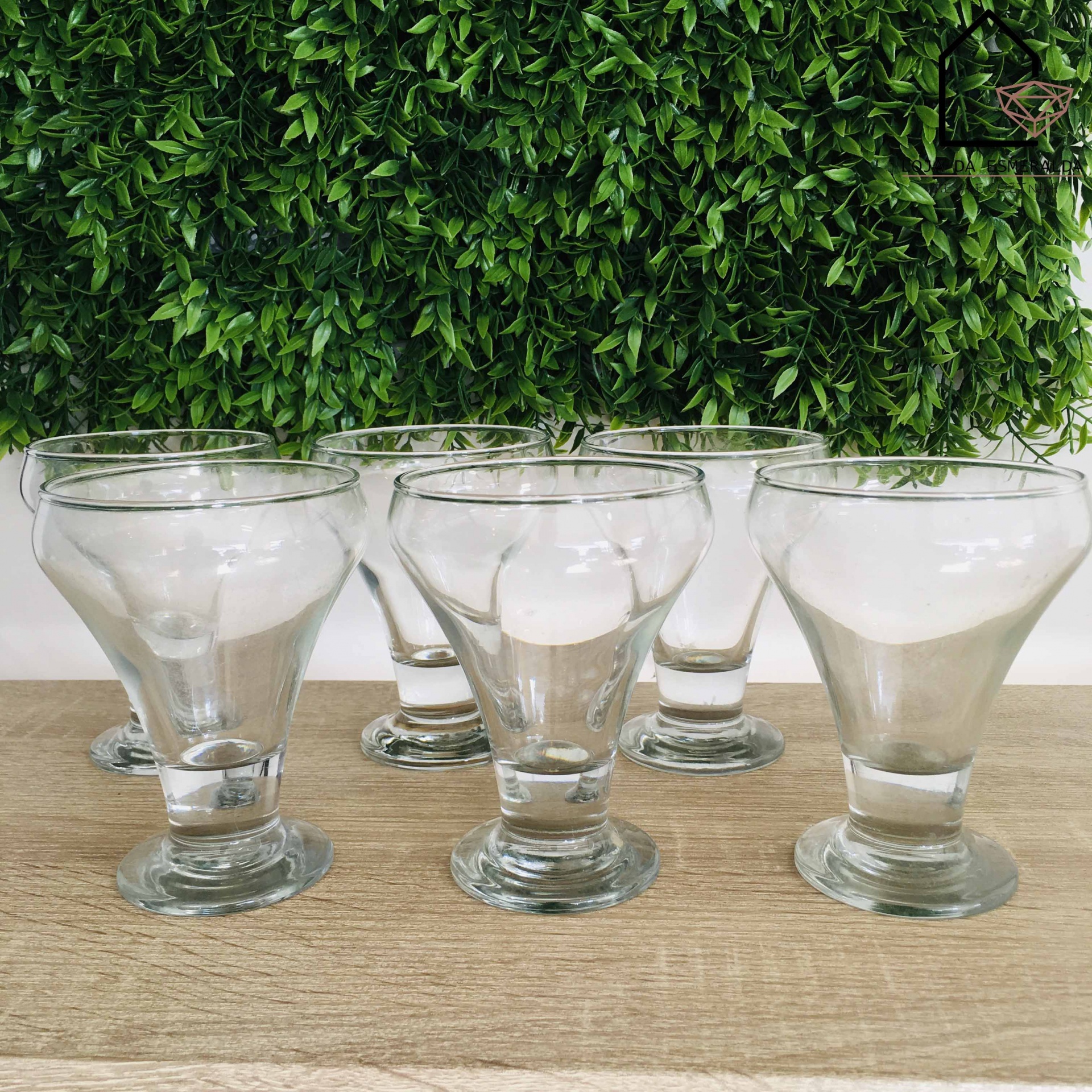 Seis copos de vidro transparente sobre mesa de madeira com fundo de folhas verdes