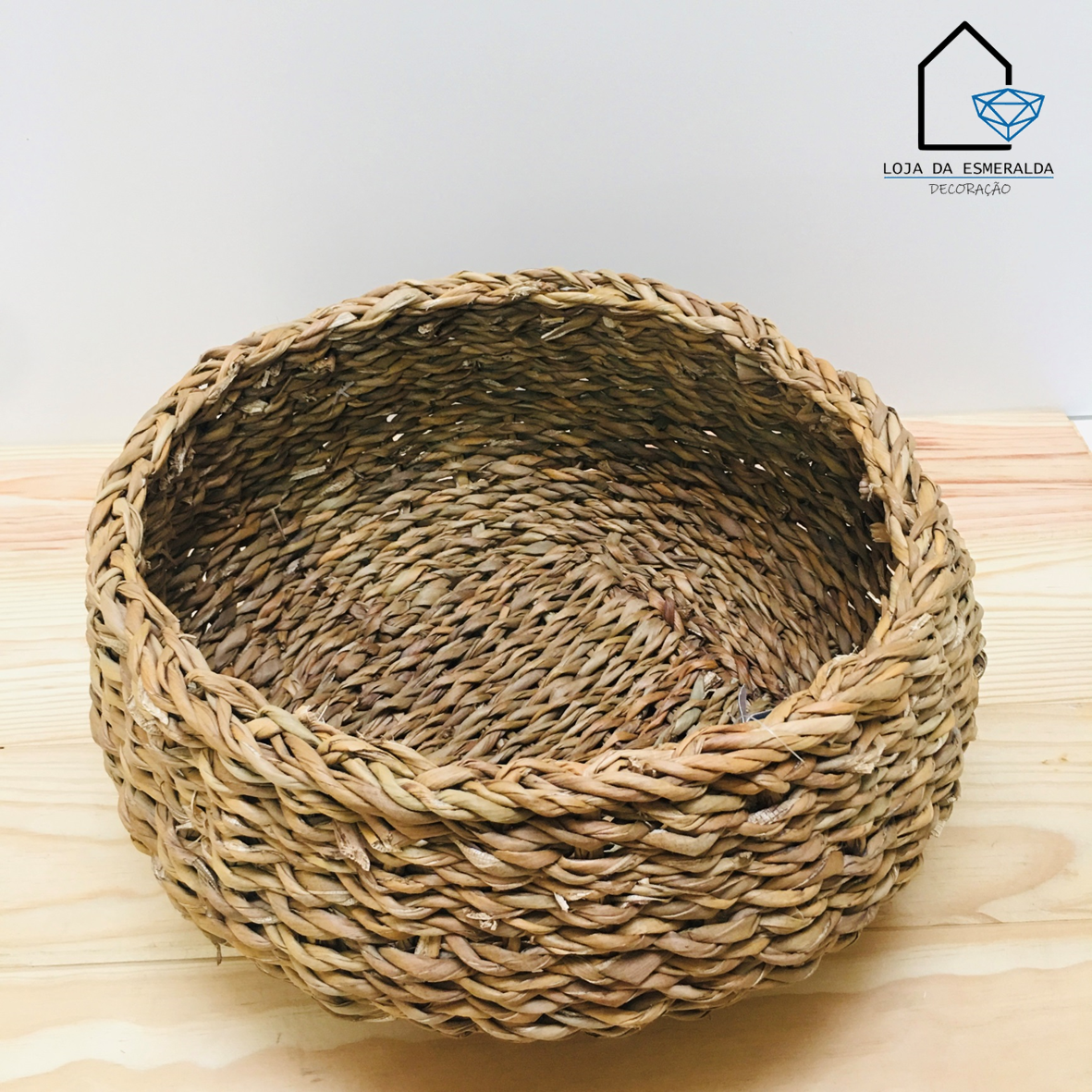 Cesto de fibra natural trançada castanho claro sobre madeira clara com logotipo