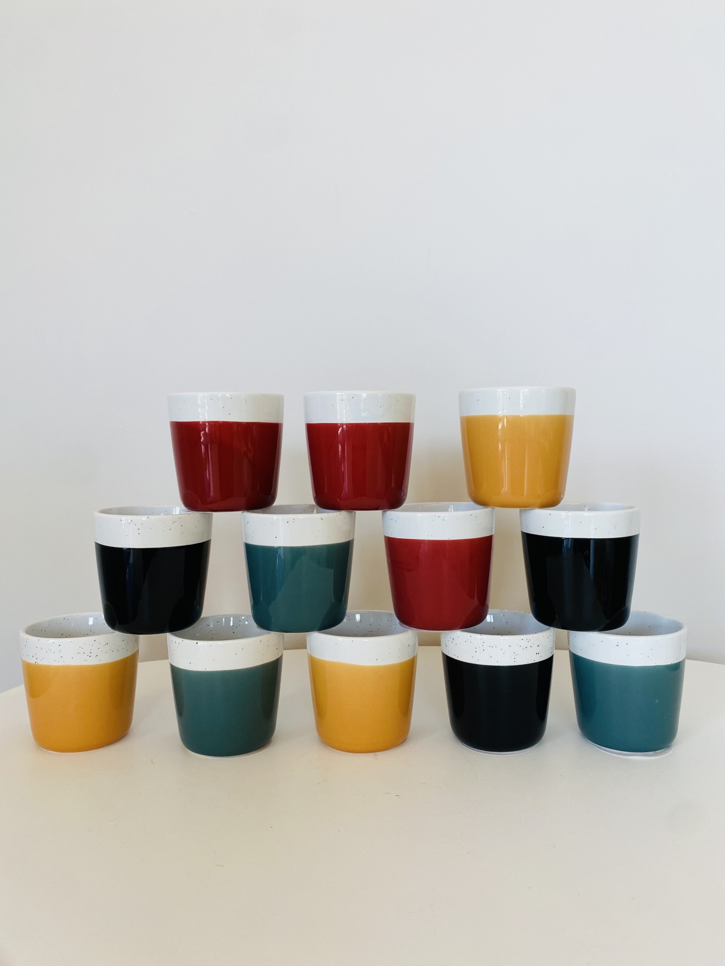 Doze copos cerâmicos coloridos empilhados em pirâmide sobre superfície branca.