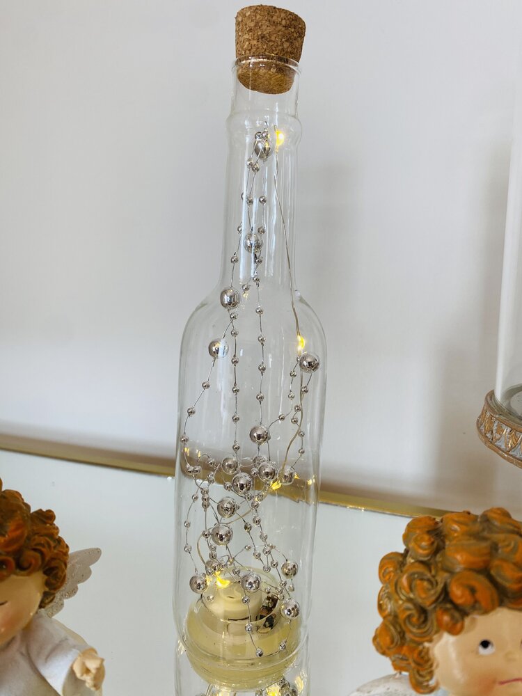 Garrafa de vidro com luzes LED e decoração prateada entre figuras angelicais