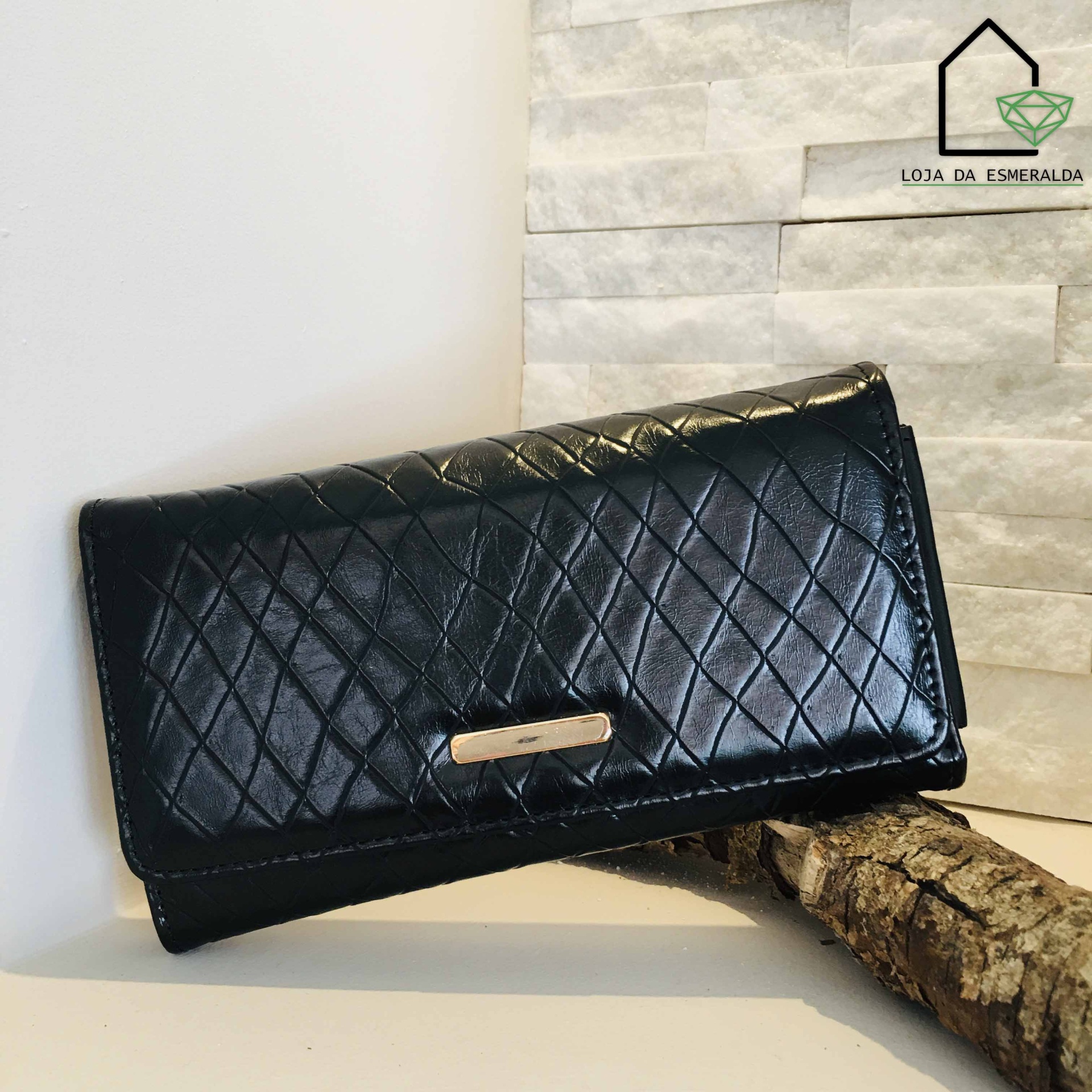 Carteira feminina preta matelassé com detalhe metálico em superfície branca e fundo de parede de pedra