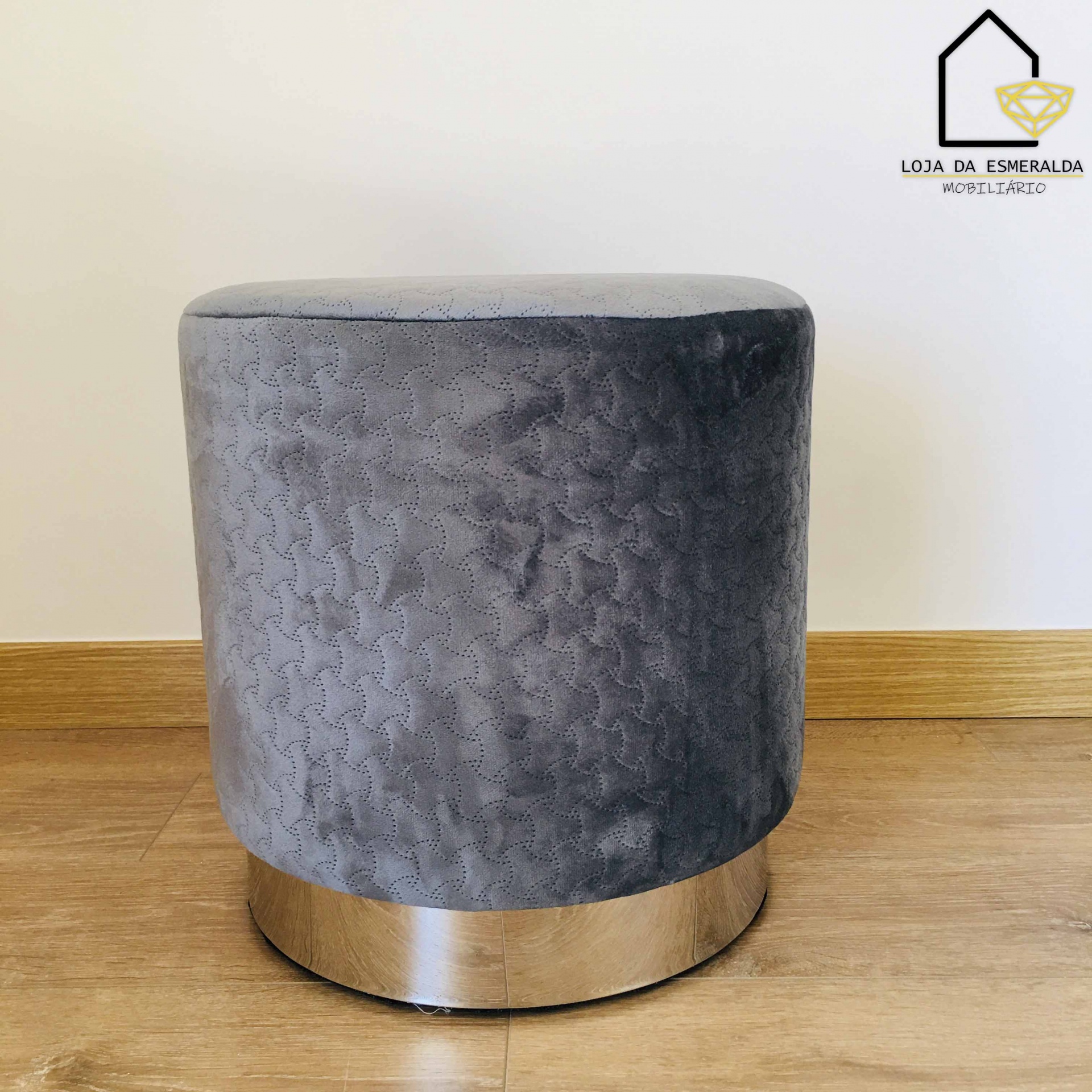 Pufe cinza com base metálica prateada sobre piso de madeira