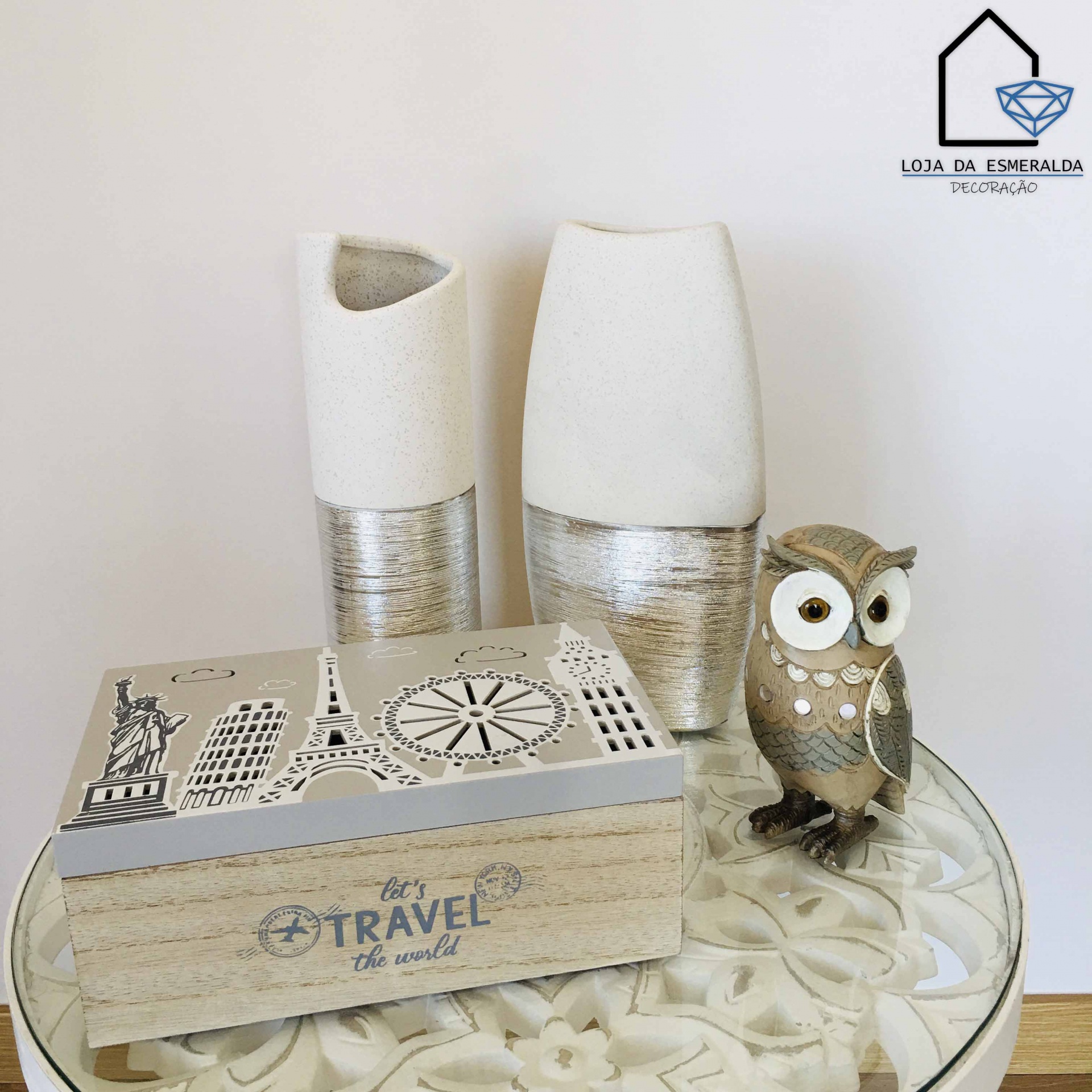 Conjunto decorativo com jarras cerâmicas, caixa de madeira Travel the world e figura de coruja numa mesa redonda de vidro