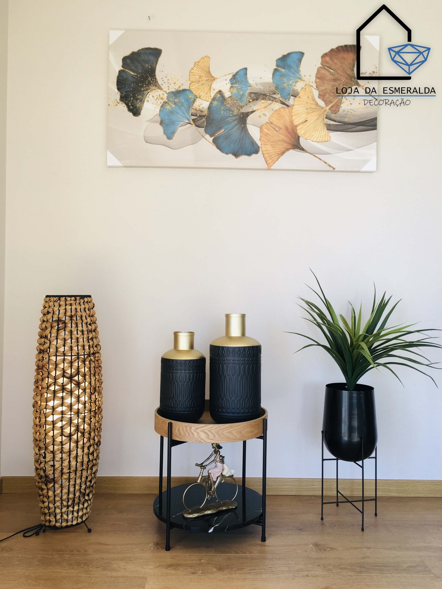 Conjunto decorativo com luminária tecida, garrafas pretas, vaso com planta e quadro de folhas coloridas