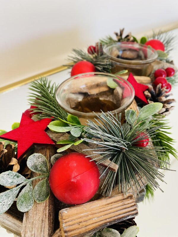 Arranjo natalício com madeira, bolas e estrelas vermelhas, ramos verdes artificiais e suporte de vela de vidro.