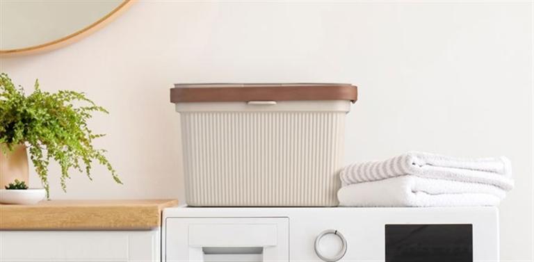 Caixa de arrumação plástica cinza com tampa castanha na máquina de lavar roupa branca com toalhas brancas e planta verde