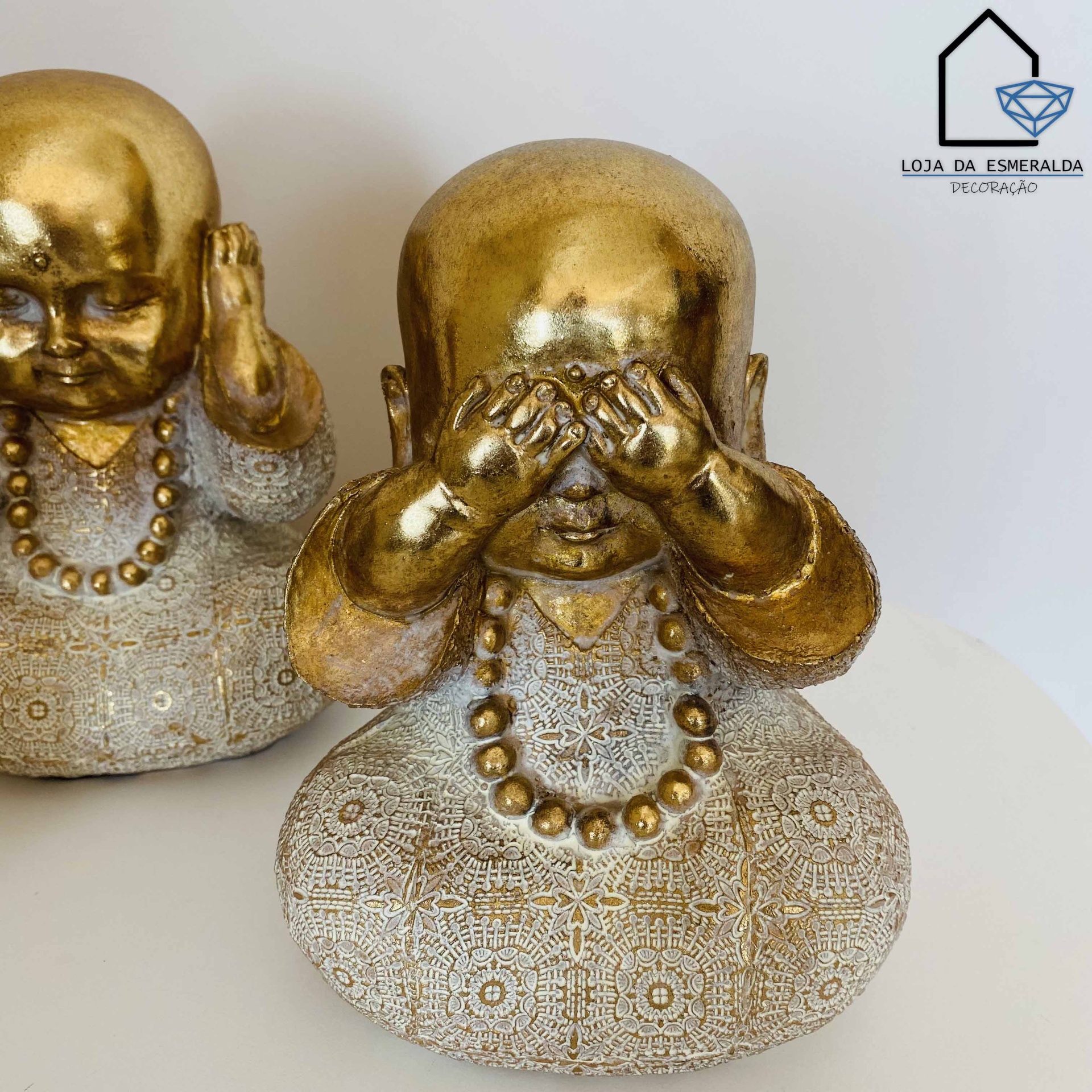 Estatuetas de buda douradas e cinza com padrão geométrico e contas, em fundo branco