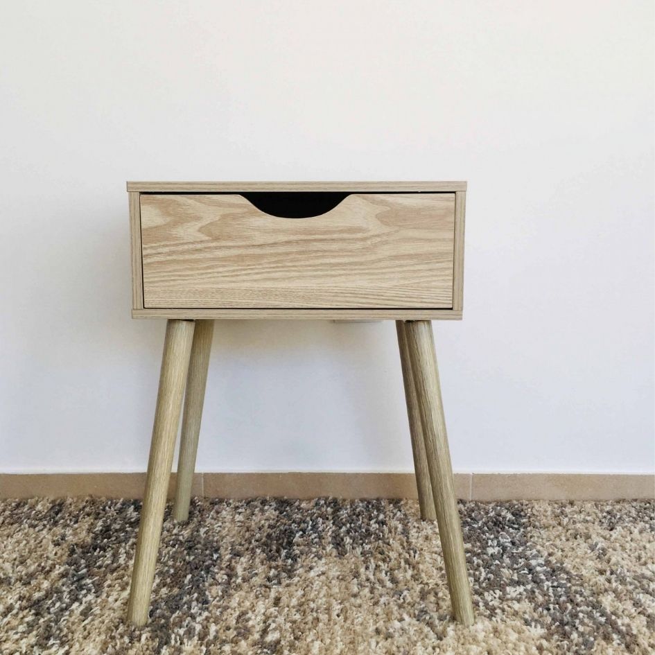Móvel de pé em madeira clara com uma gaveta e pernas cilíndricas inclinadas sobre tapete.
