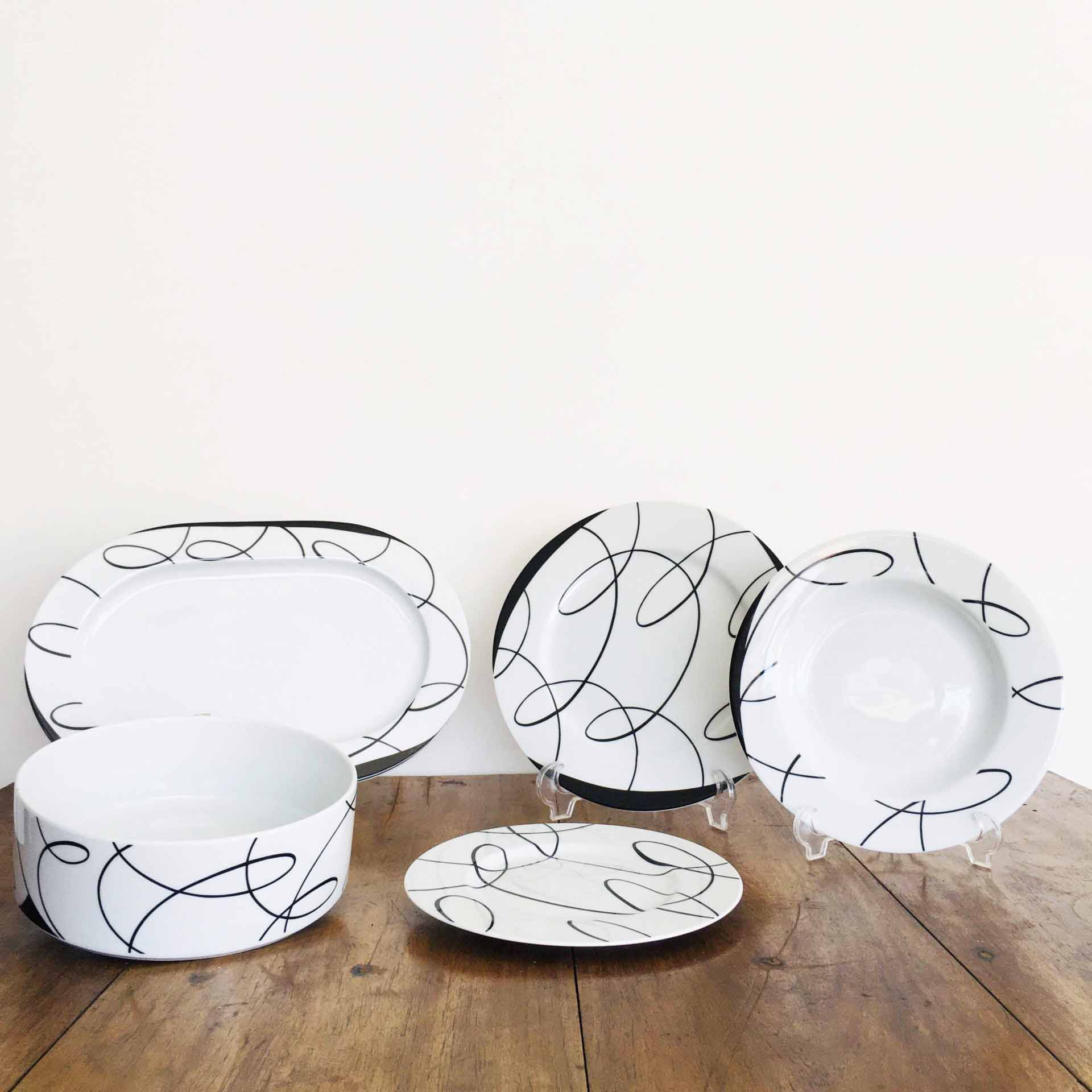 Conjunto de loiça branca com linhas pretas curvas, vários pratos e travessa sobre mesa de madeira.