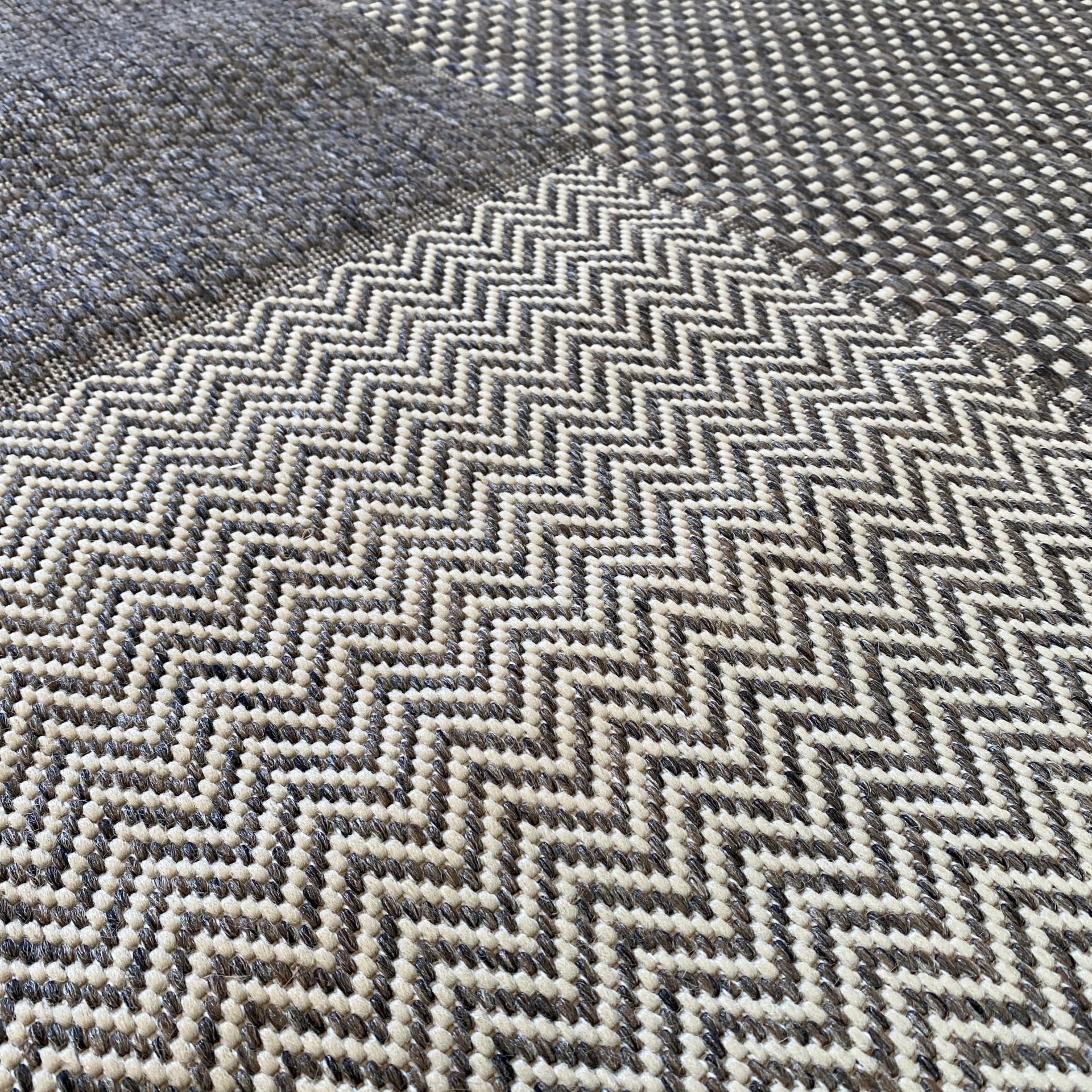 Tapete têxtil com padrão em chevron e quadrados em tons de bege, cinzento e preto.