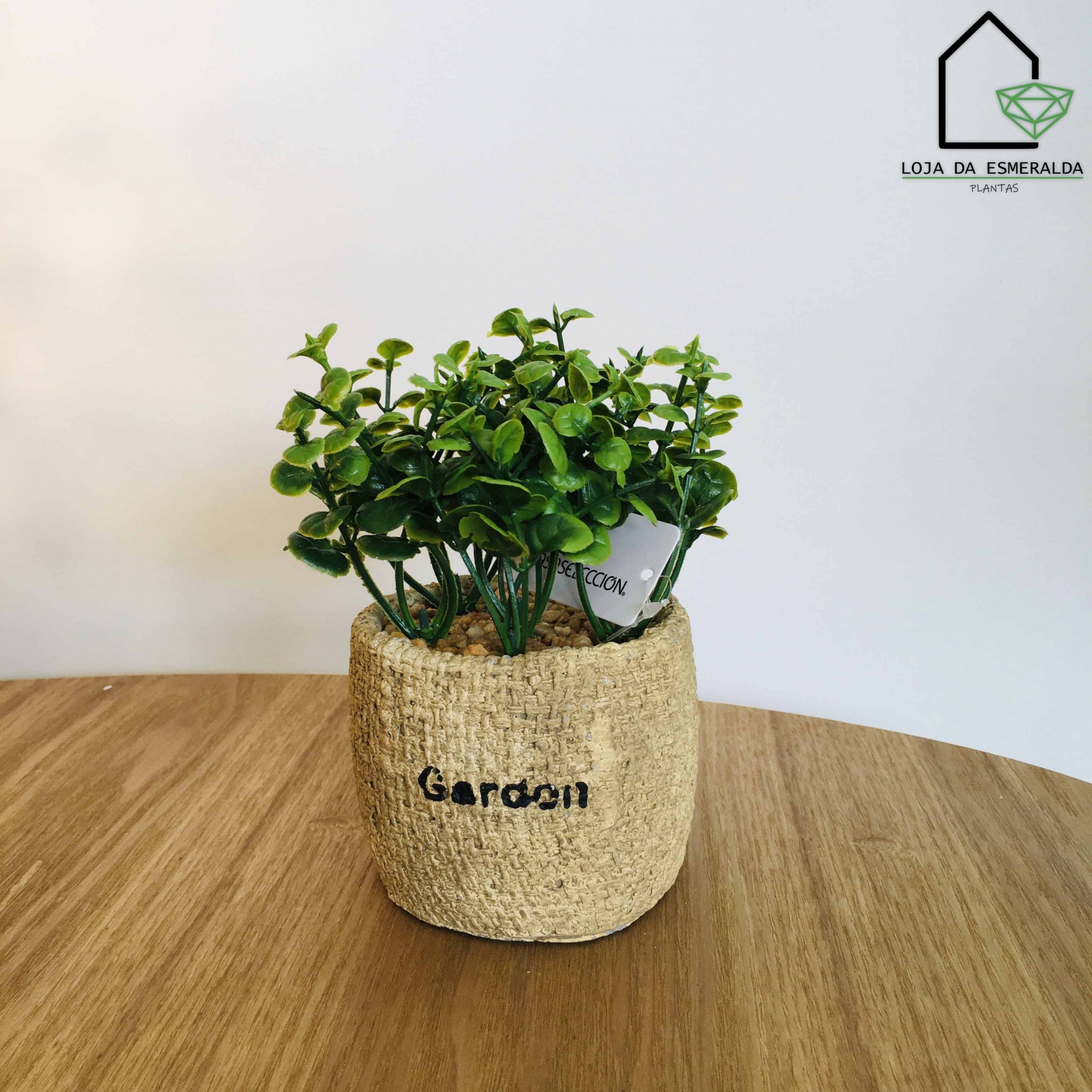 Vaso de planta com suporte de tecido bege e planta verde artificial sobre mesa de madeira