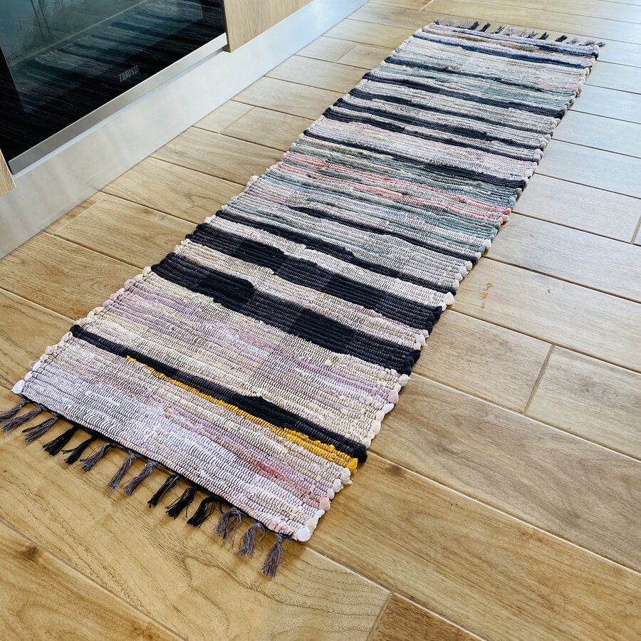 Tapete estreito e longo com padrão de riscas coloridas e franjas, sobre chão de madeira clara