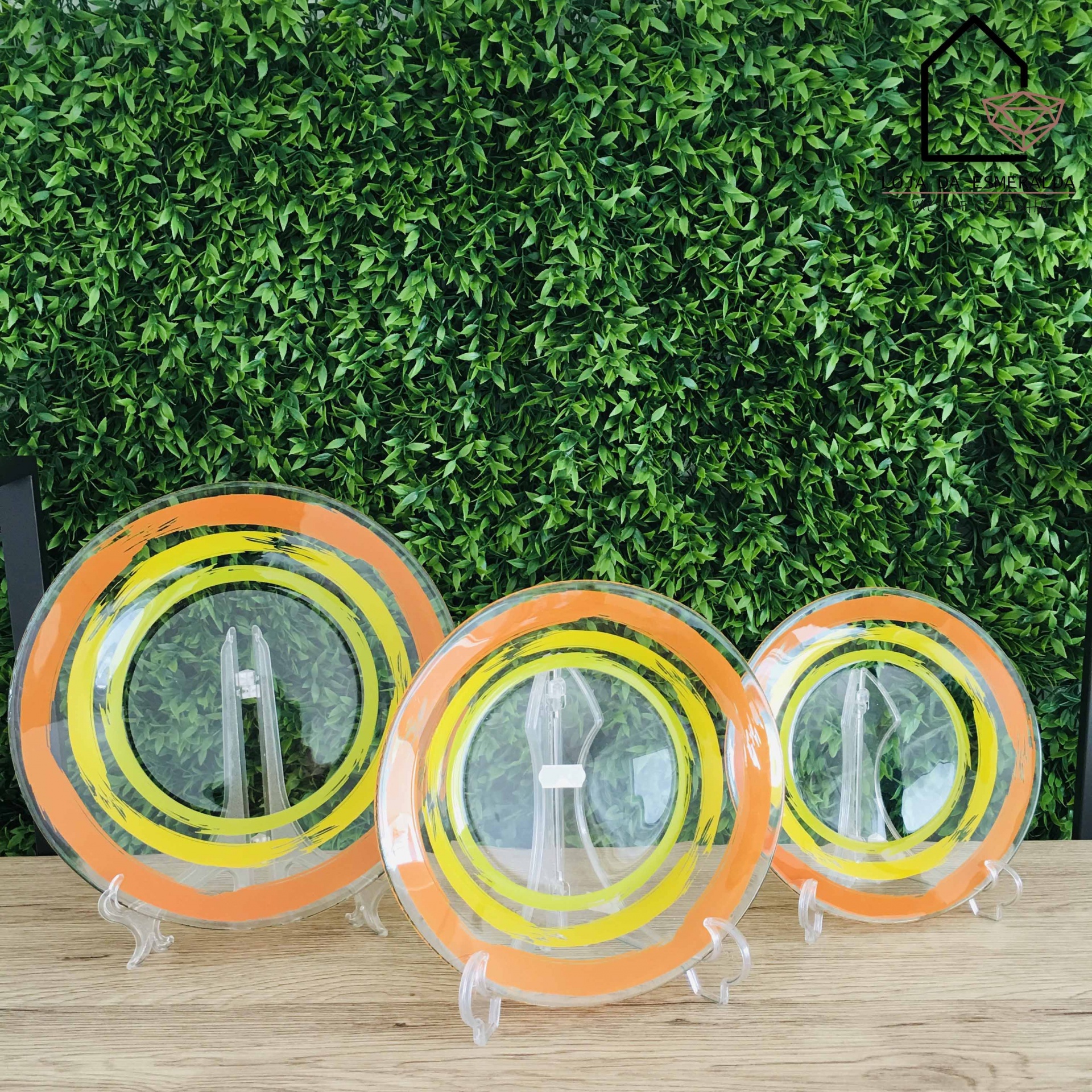 Três pratos de vidro com padrões coloridos em suporte transparente sobre mesa de madeira clara