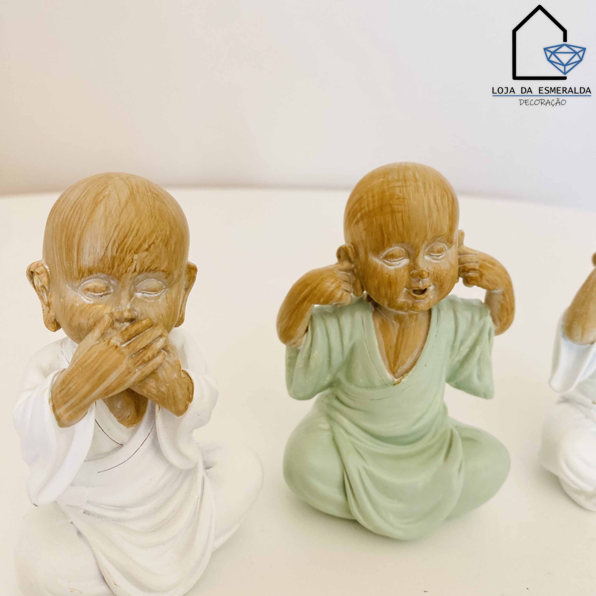Três estatuetas de crianças em túnicas, em estilo monge, sobre superfície branca com logotipo LOJA DA ESMERALDA