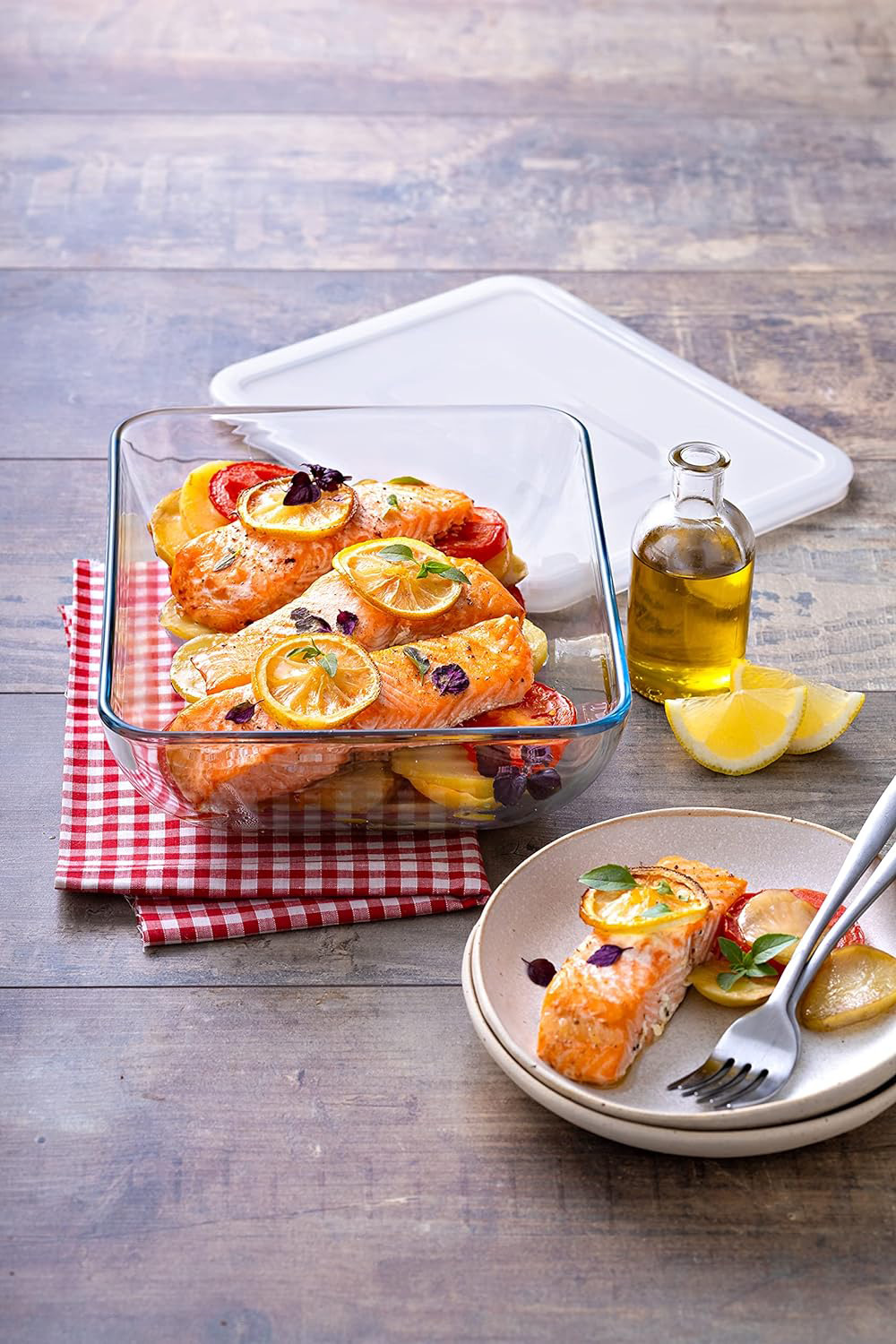 Travessa de vidro com salmão e rodelas de limão e tomate, prato com porção da comida, garrafa de óleo e fatias de limão sobre mesa de madeira.