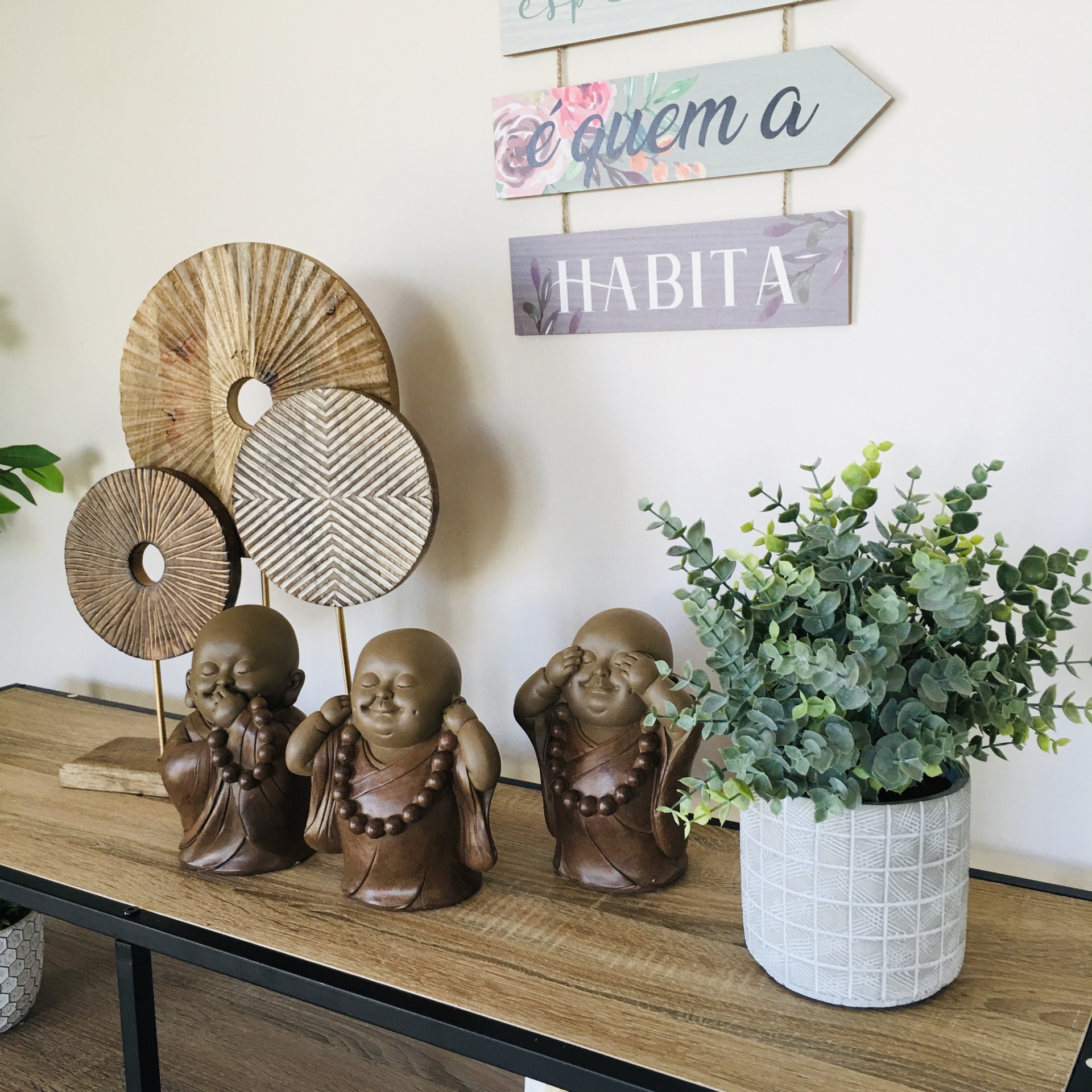 Estatuetas de madeira, decorações circulares, planta em jarra branca e placas decorativas com texto na parede