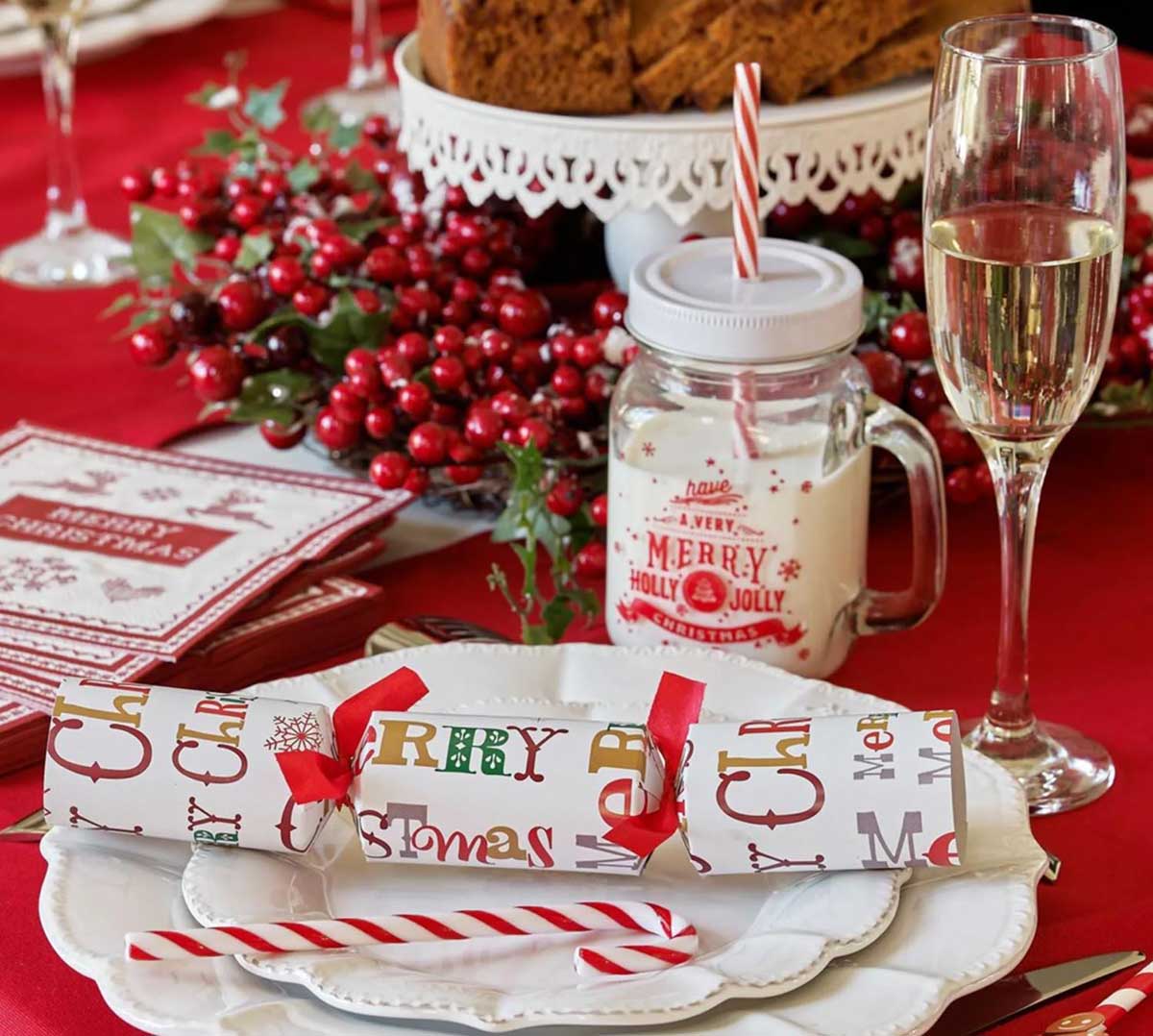 Conjunto de mesa decorada para Natal com prato branco, embalagem de papel colorido, pau de doce, copo de vidro e frasco com bebida sobre toalha vermelha.