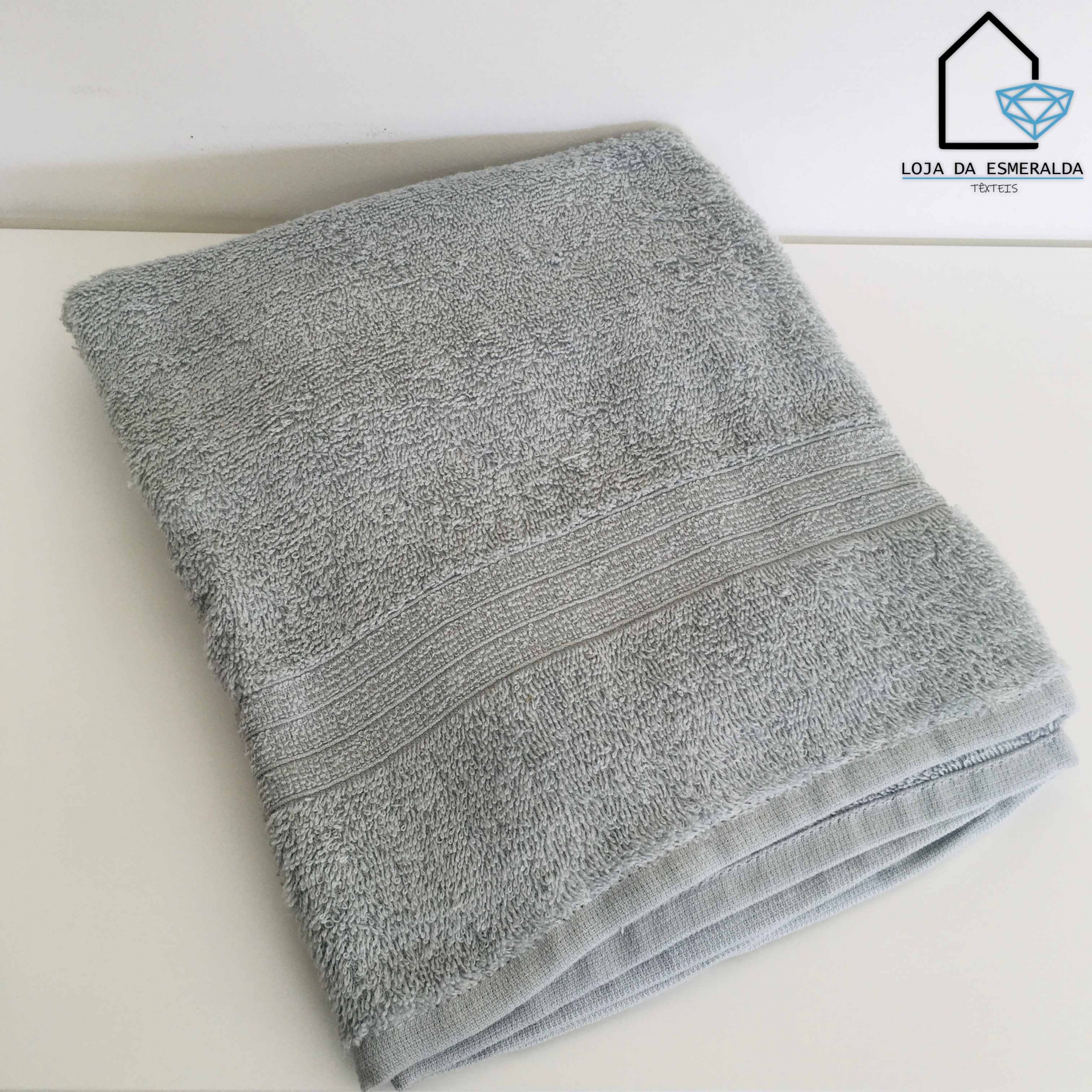 Toalha cinzenta dobrada sobre superfície branca com logótipo 'LOJA DA ESMERALDA'