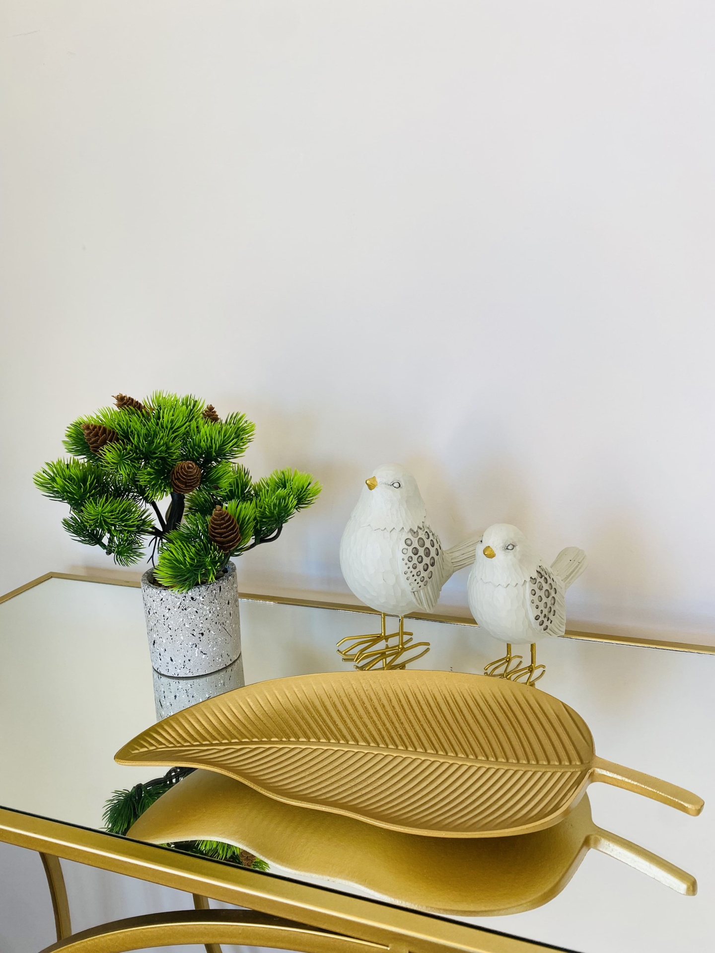 Decorativo folha dourada, pássaros brancos e planta verde em vaso cinza sobre mesa espelhada.