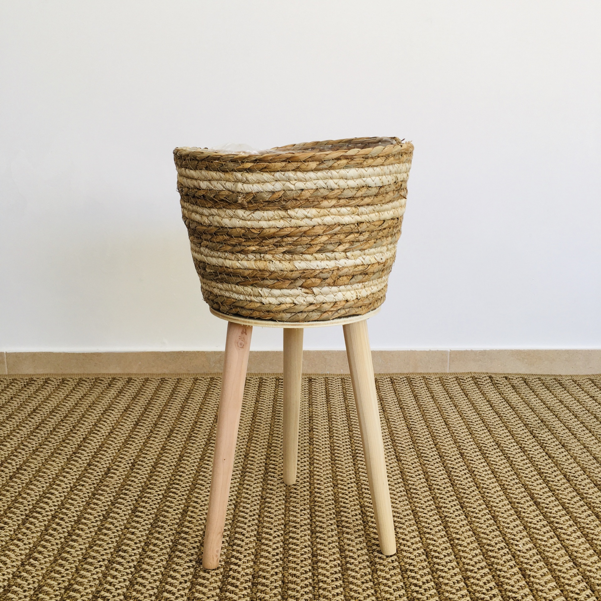 Suporte para plantas com vaso de vime trançado em tons castanhos e bege, base de madeira clara.