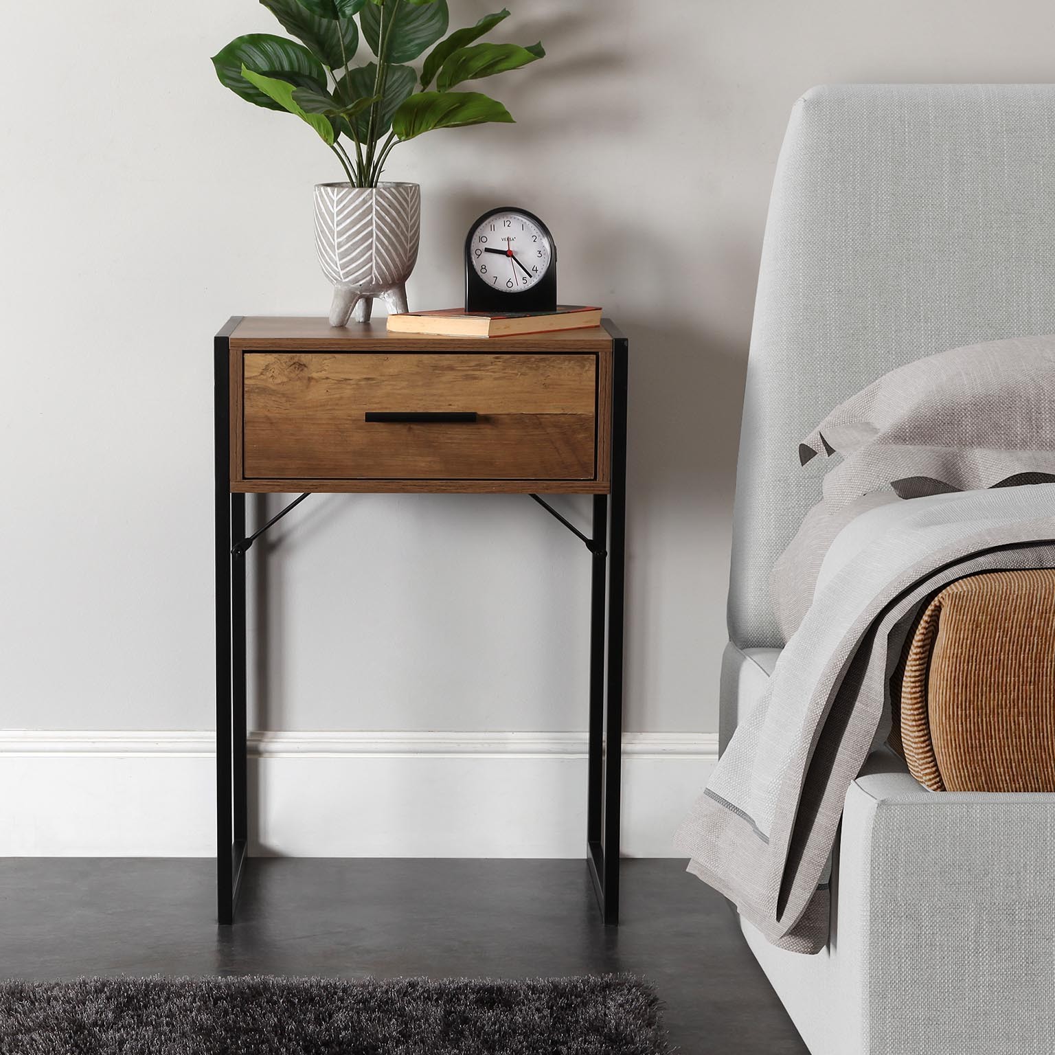 Mesa de cabeceira em madeira com vaso e relógio ao lado de cama cinzenta