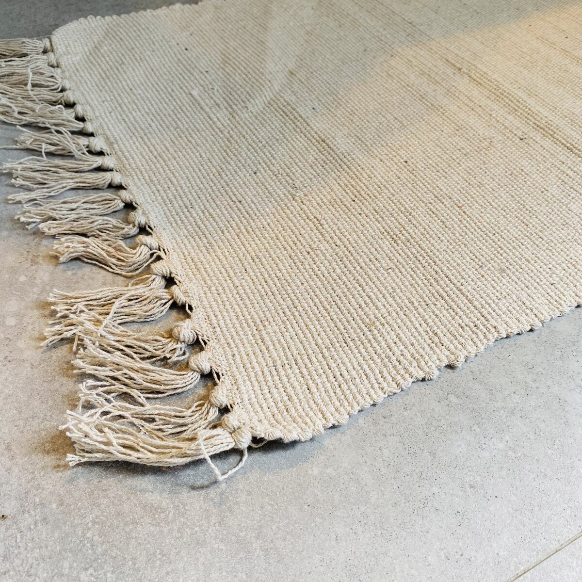 Tapete bege claro com franjas e textura tecida sobre chão de cimento cinzento