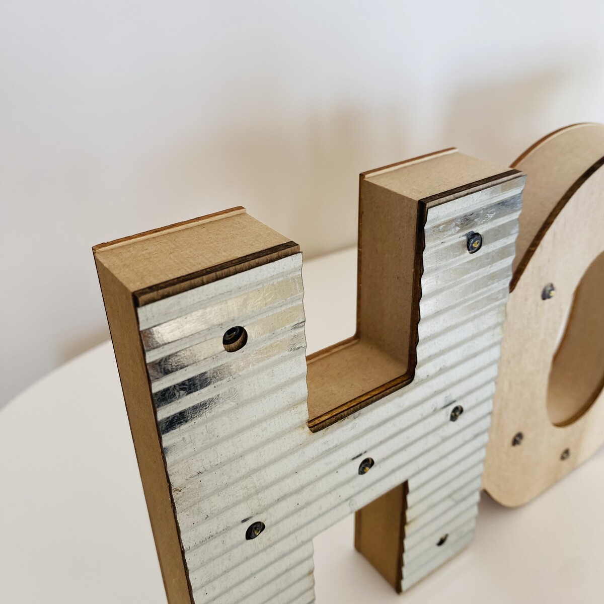 Letra decorativa 'H' em madeira com frente metálica prateada ondulada.