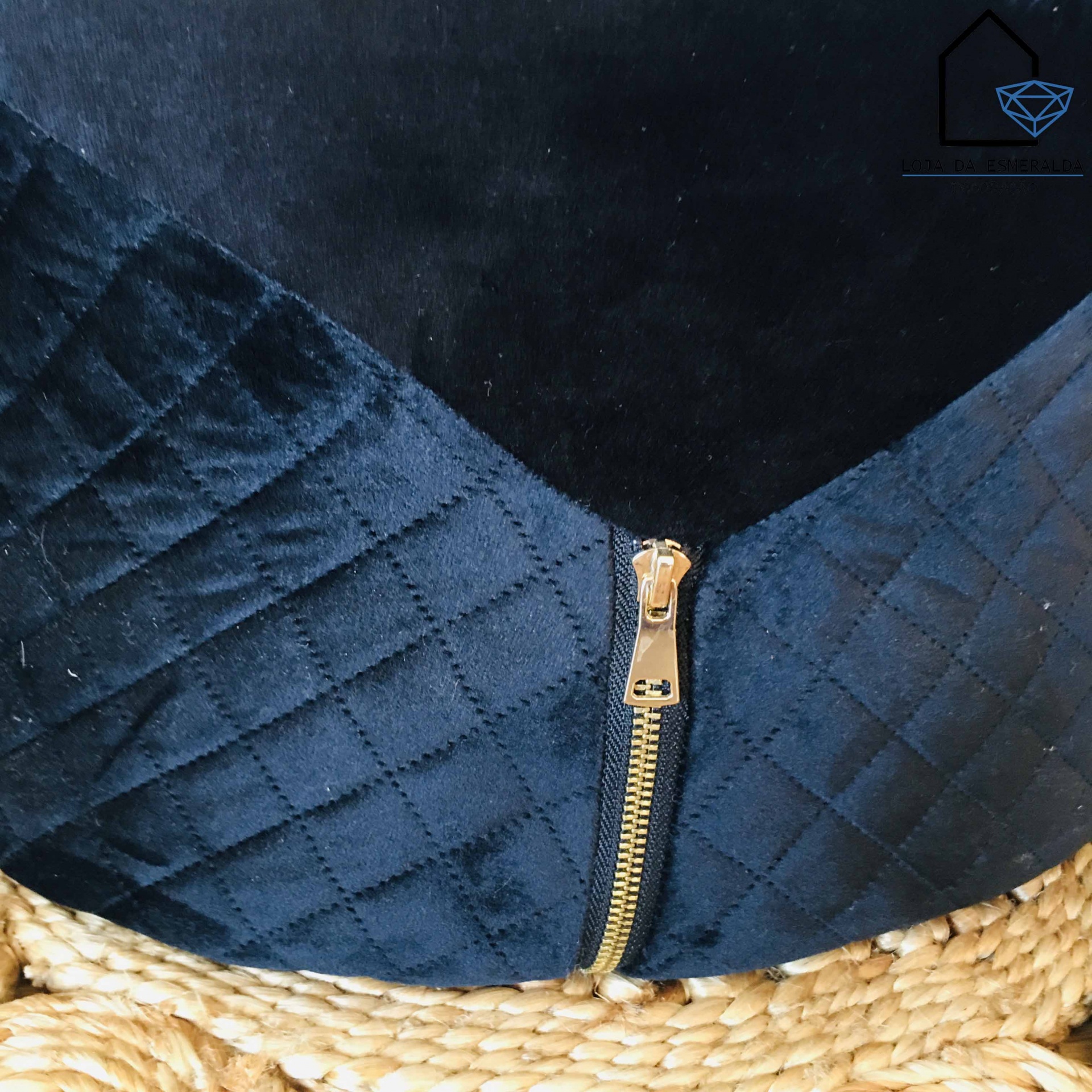 Peça de roupa escura com padrão em losangos e fecho de correr dourado sobre superfície trançada