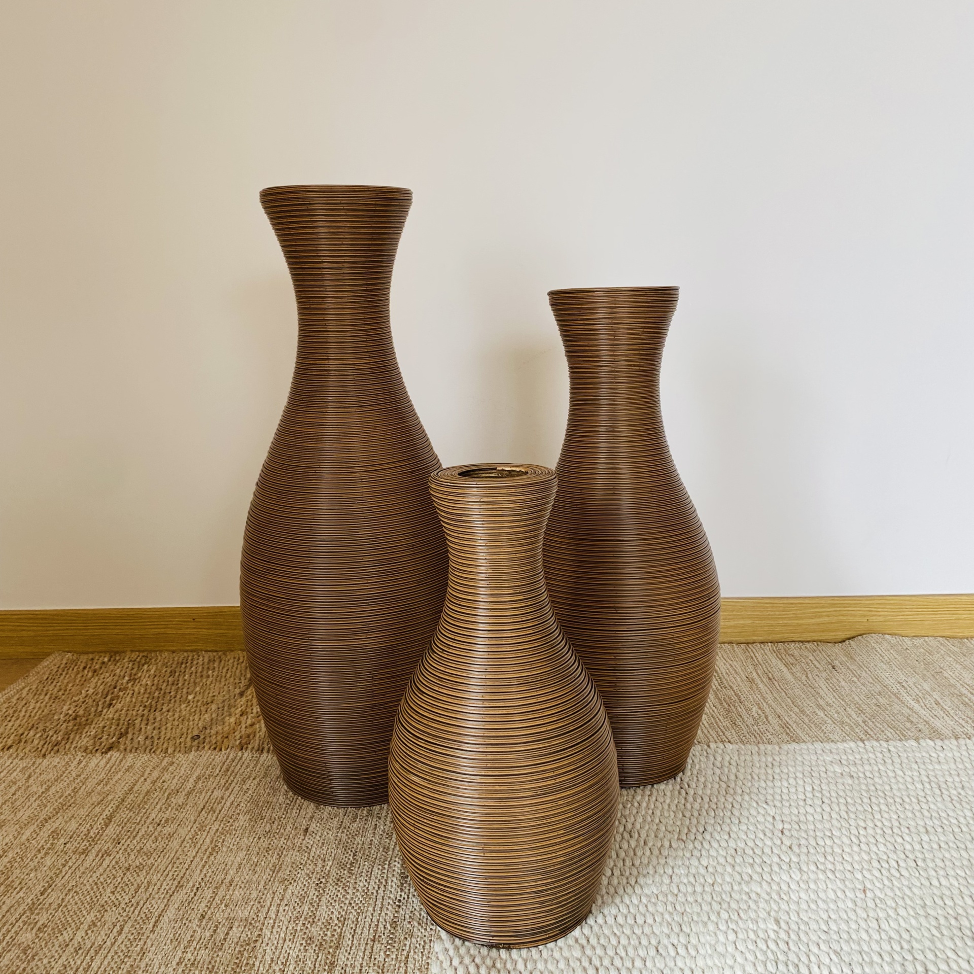 Três vasos decorativos de rattan marrom sobre tapetes bege e castanho com parede branca ao fundo