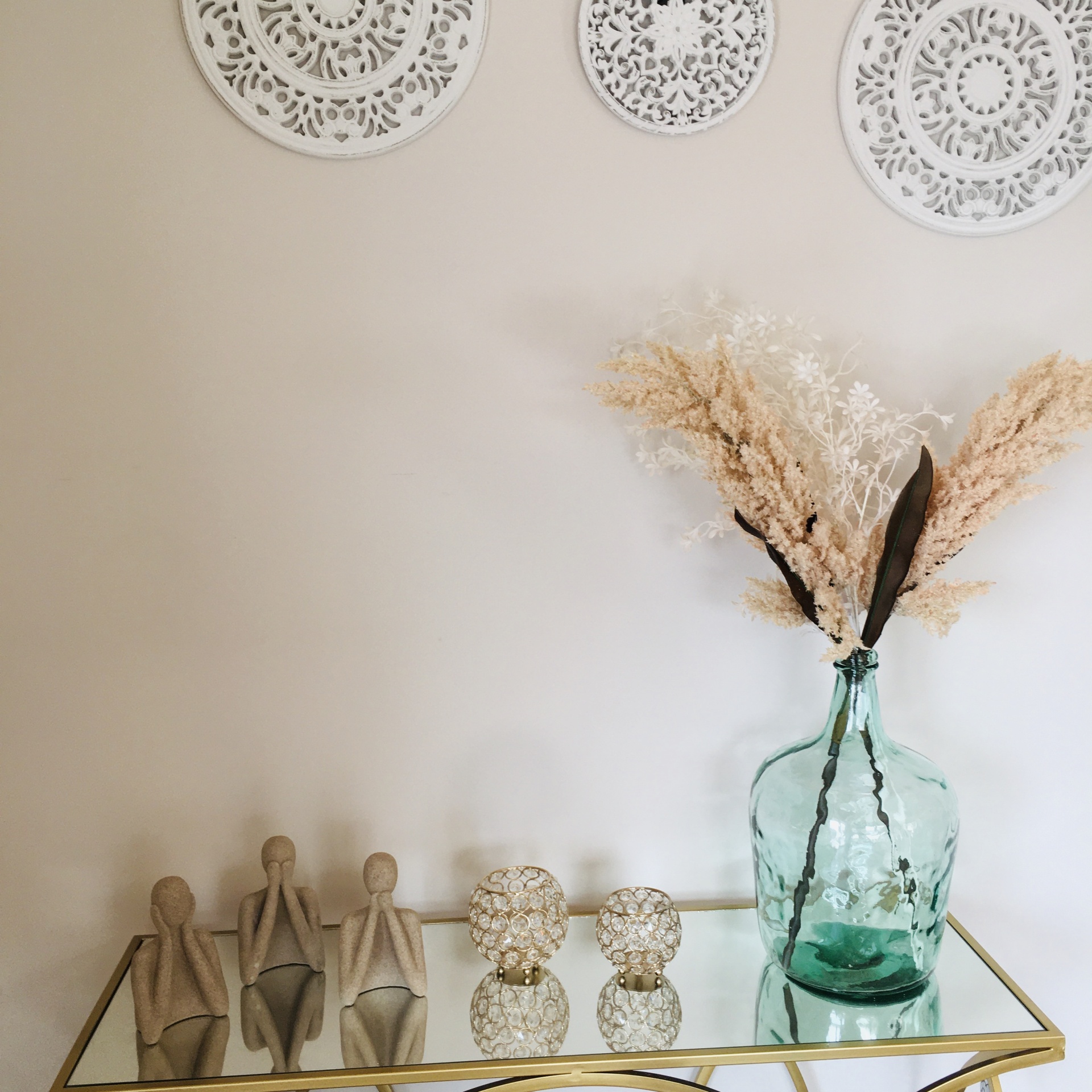 Mesa de vidro com base dourada com três estatuetas de figuras humanas bege, dois castiçais dourados e vaso verde com ramos secos