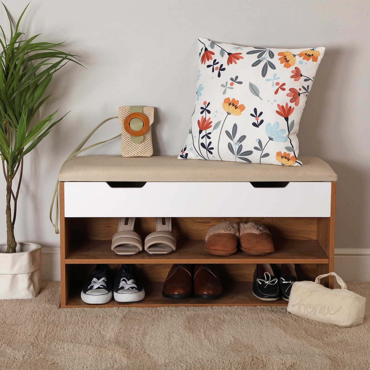 Banco organizador com assento almofadado bege, calçado, almofada floral e planta