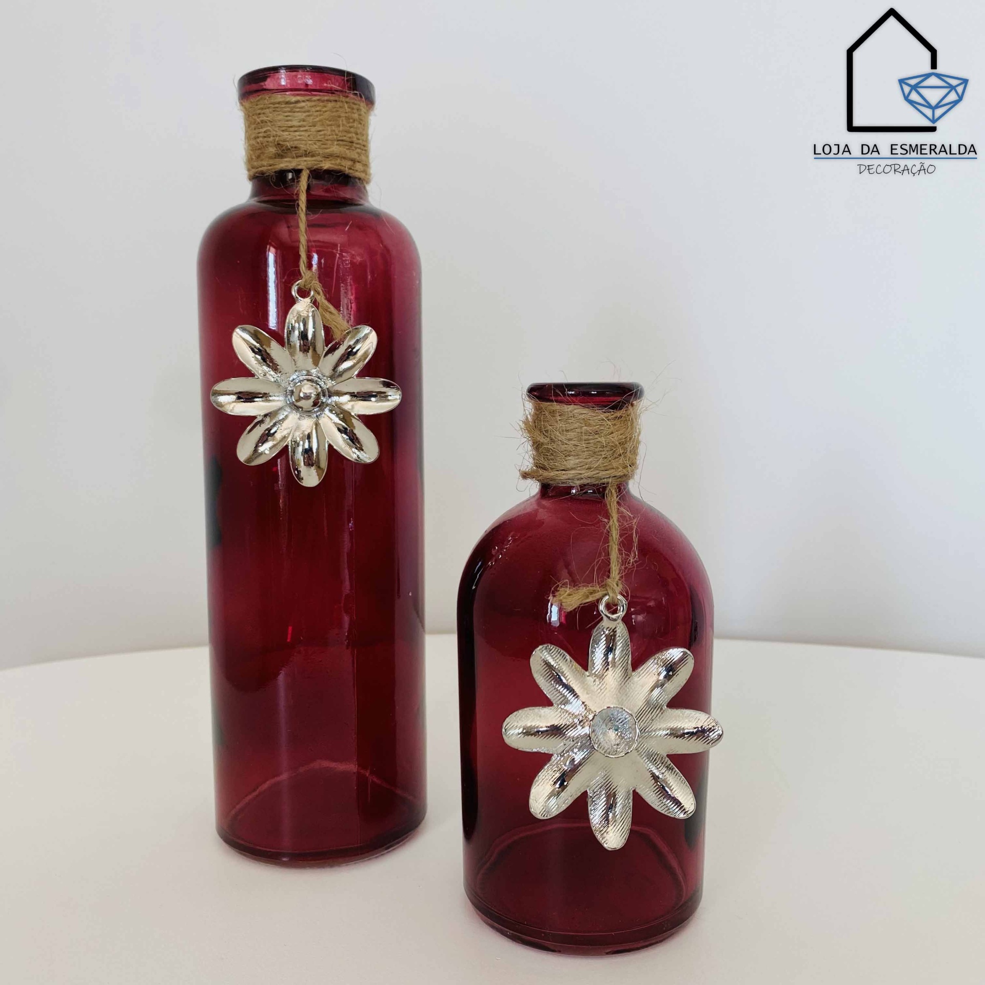 Dois frascos decorativos vermelhos com pendentes florais prateados e corda no gargalo sobre superfície branca