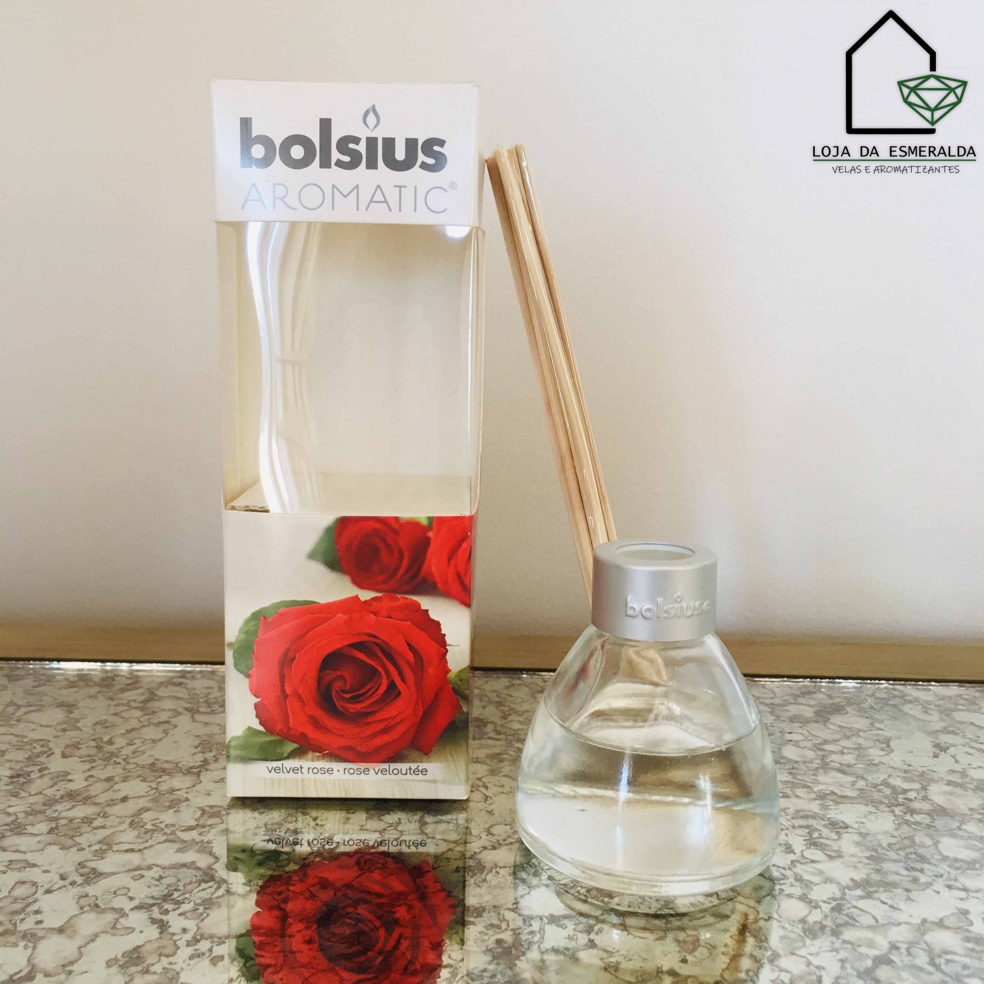 Difusor de aroma com frasco de vidro e pauzinhos de madeira ao lado de embalagem com imagem de rosa vermelha