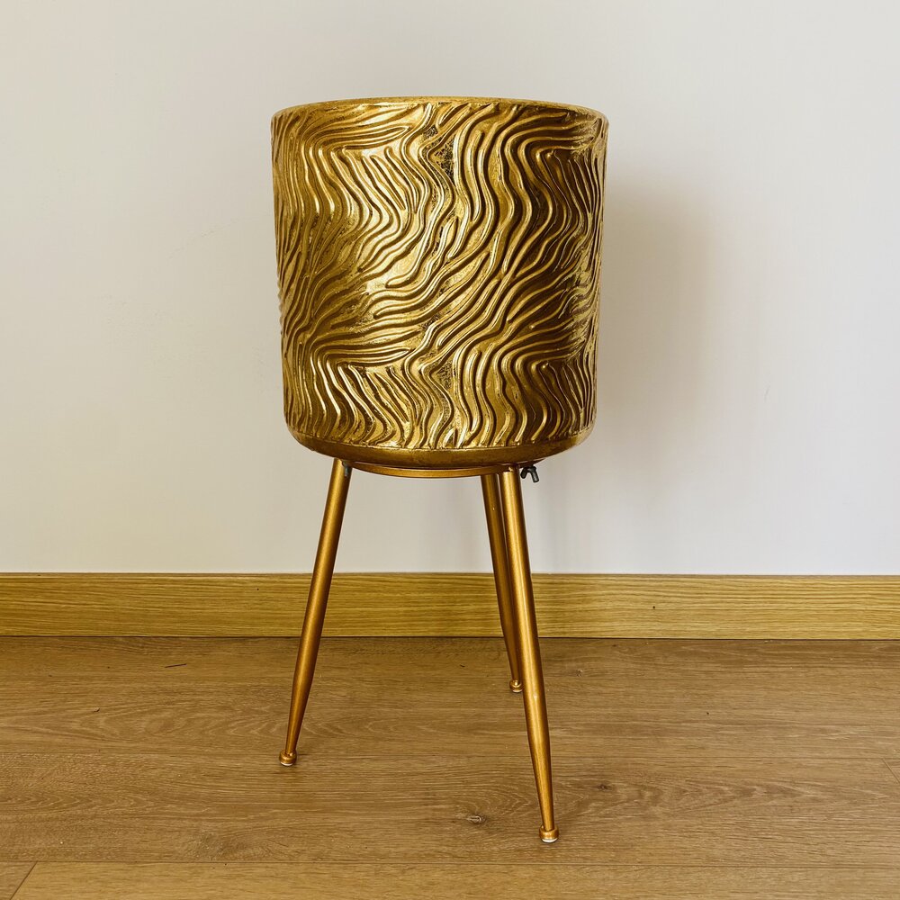 Vaso dourado texturizado em suporte metálico de três pernas sobre chão de madeira