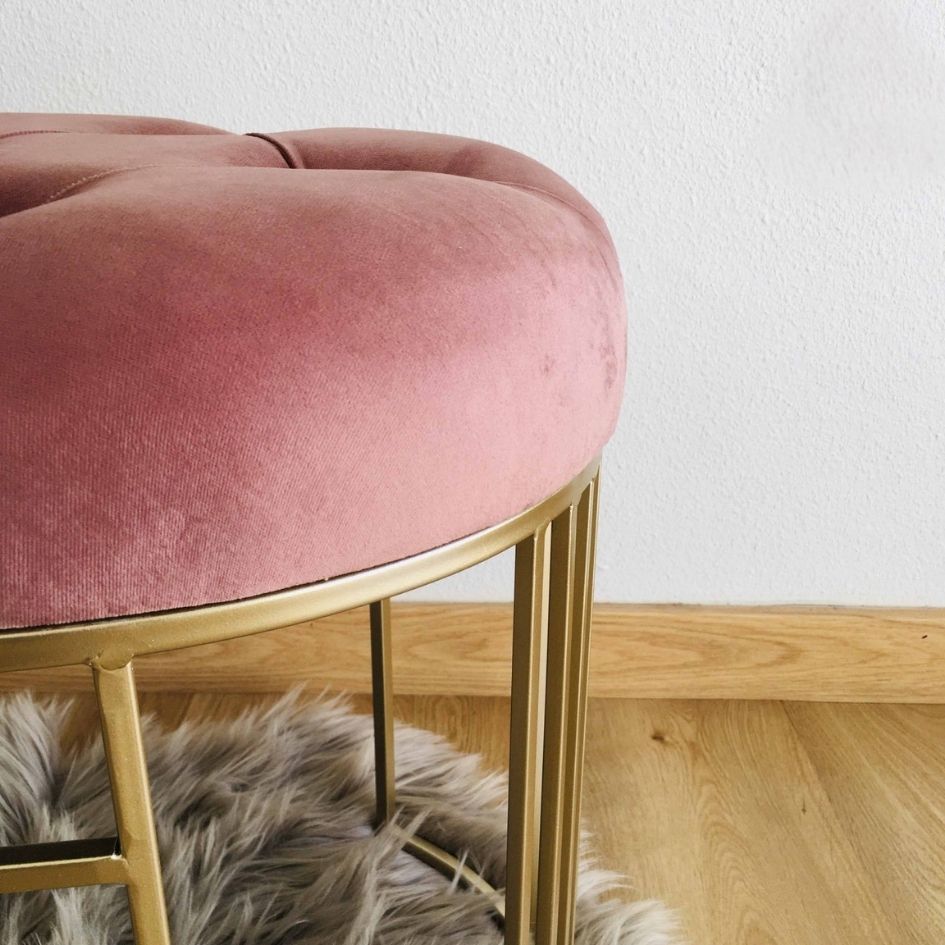 Banco redondo rosa de veludo com pernas douradas em tapete cinza no chão de madeira