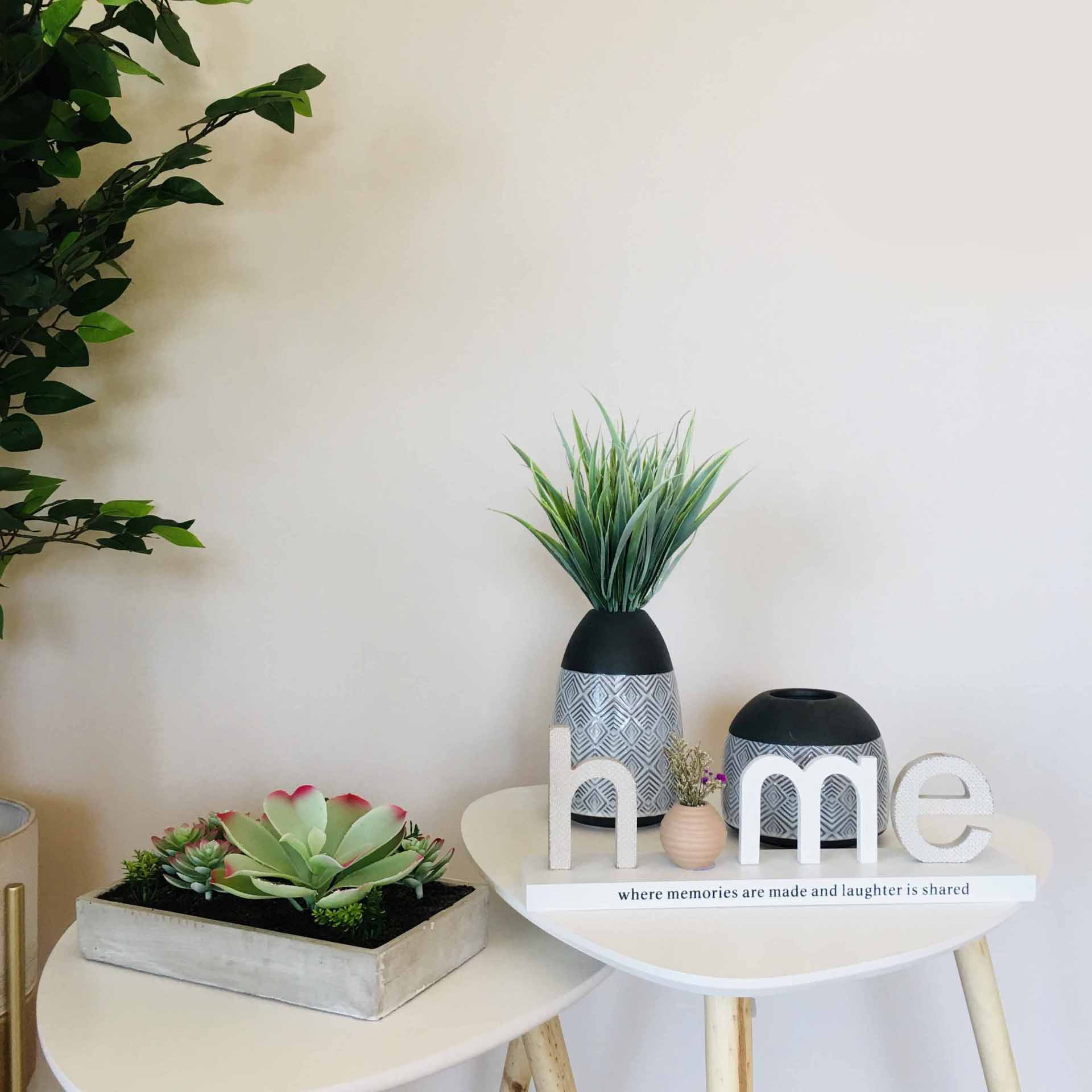 Mesa decorativa com vasos cinzas, letras 'home' brancas e plantas verdes sobre duas mesas brancas com pernas de madeira