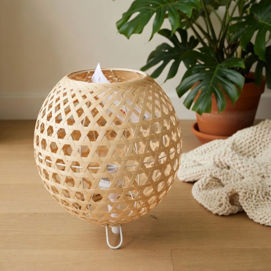 Abajur redondo de bambu trançado com base branca ao lado de planta em vaso e manta