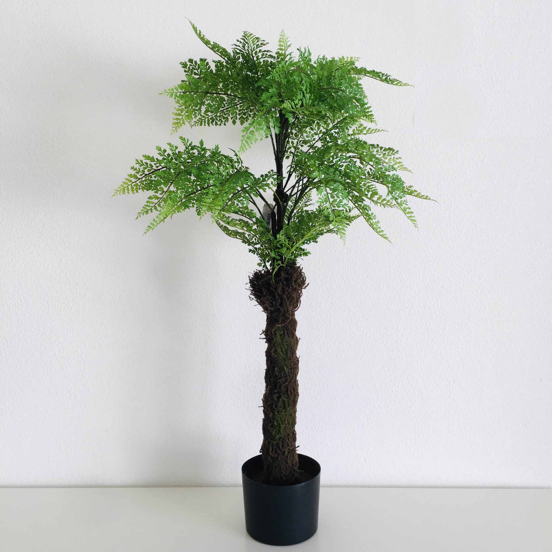 Planta artificial de samambaia em vaso preto sobre superfície branca