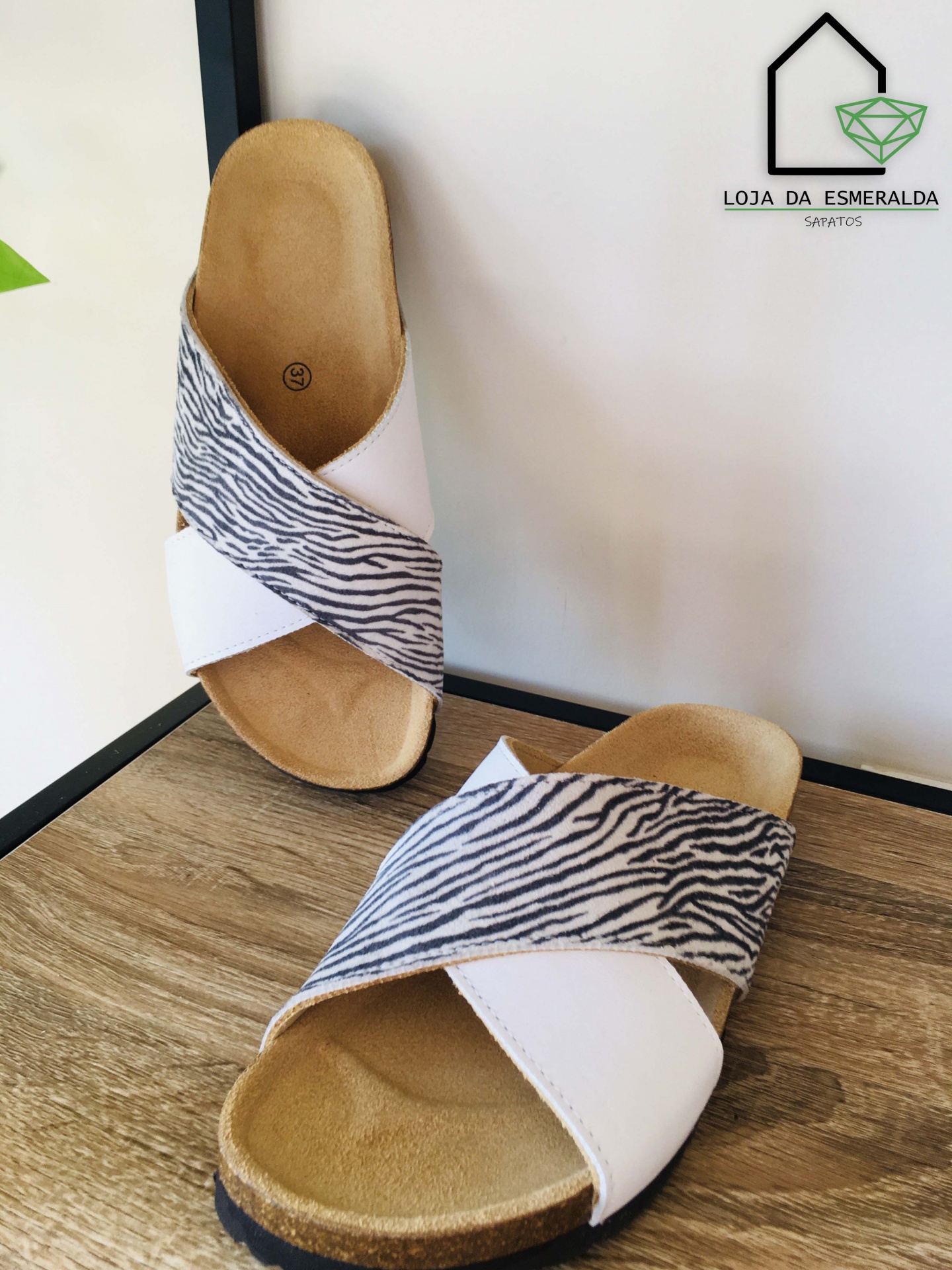 Chinelos com tiras brancas e padrão riscado preto e branco, sobre fundo de madeira com parede branca ao fundo