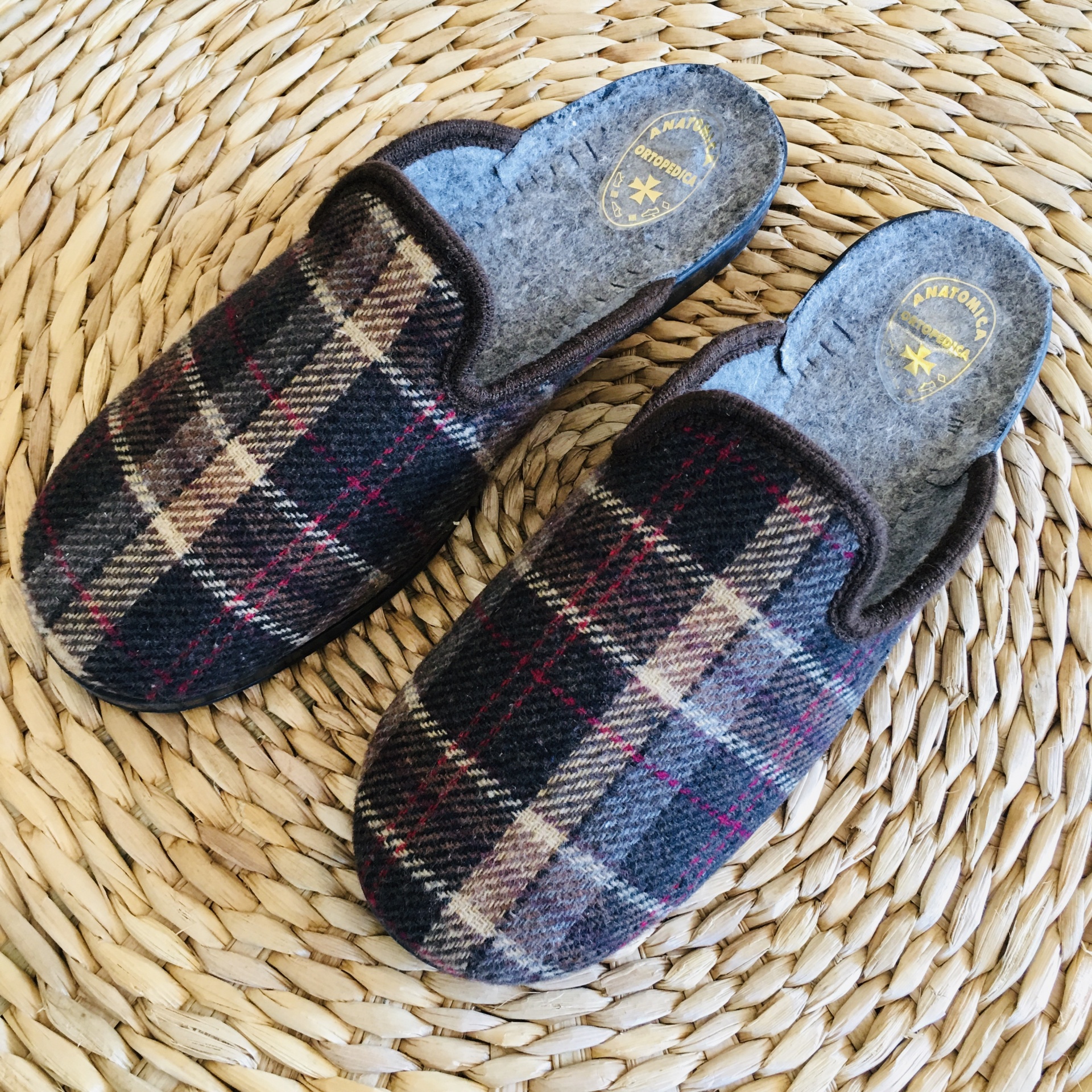 Chinelos com padrão xadrez em tons de preto, bege e vermelho, interior cinzento, sobre superfície trançada bege